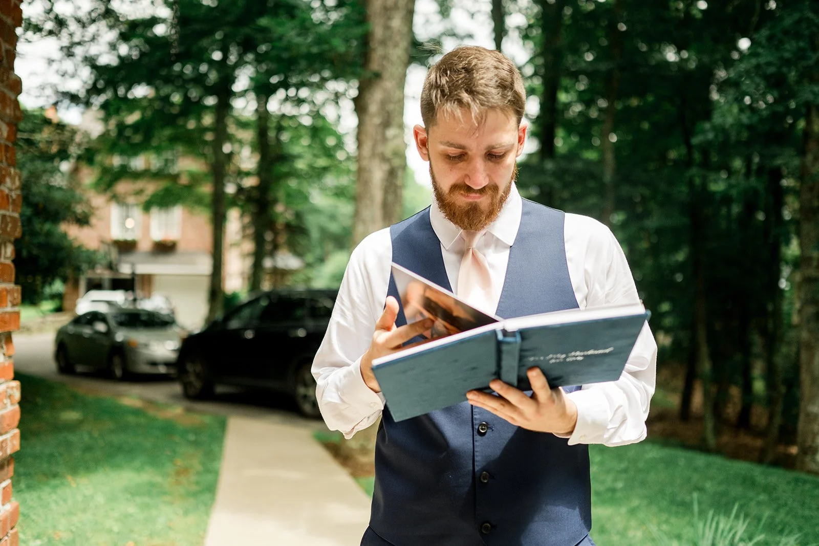 Groom reading from a blue vow book outdoors before the wedding ceremony while wearing a navy vest.