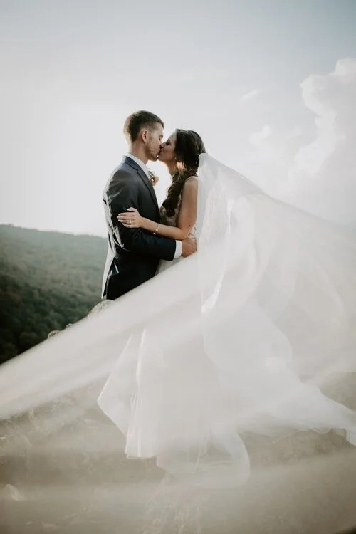 Bride and groom kissing outdoors with a long veil blowing in the wind and mountains in the background