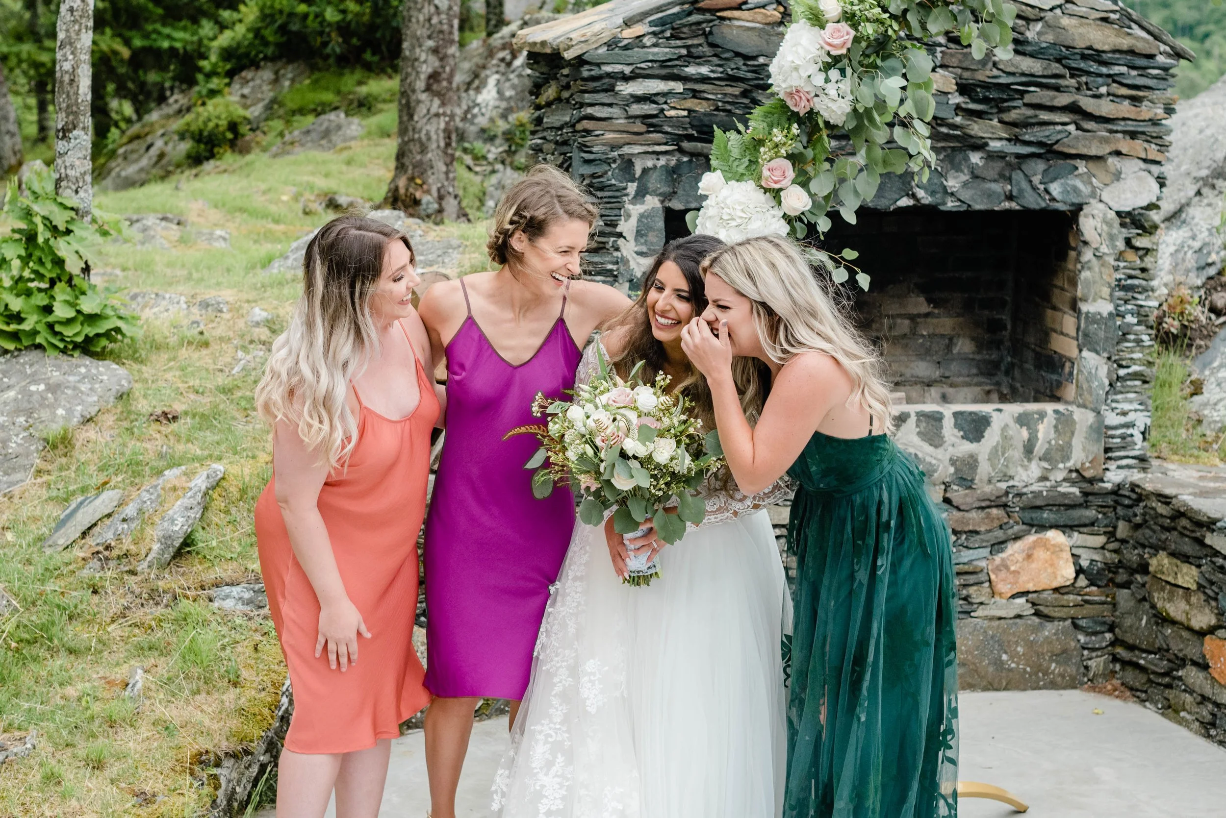 Bride laughing with bridesmaids in colorful dresses while holding a white and blush bouquet.
