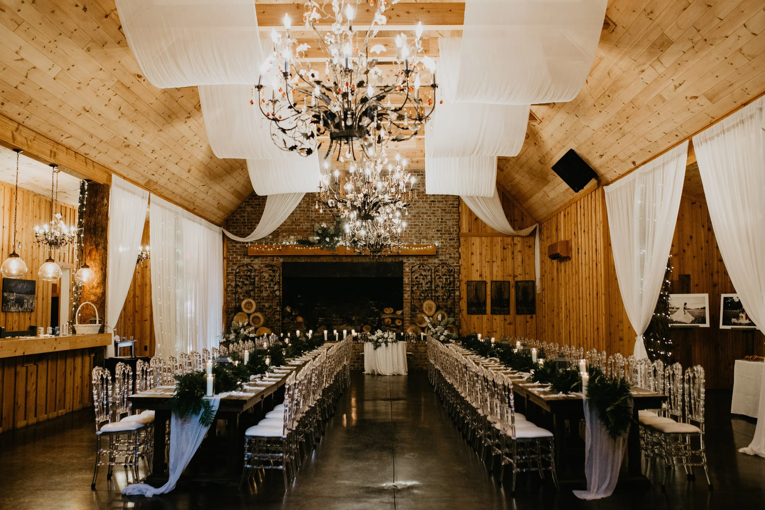 Wide view of a wooden reception hall set with long tables, clear chairs, chandeliers, and white ceiling draping