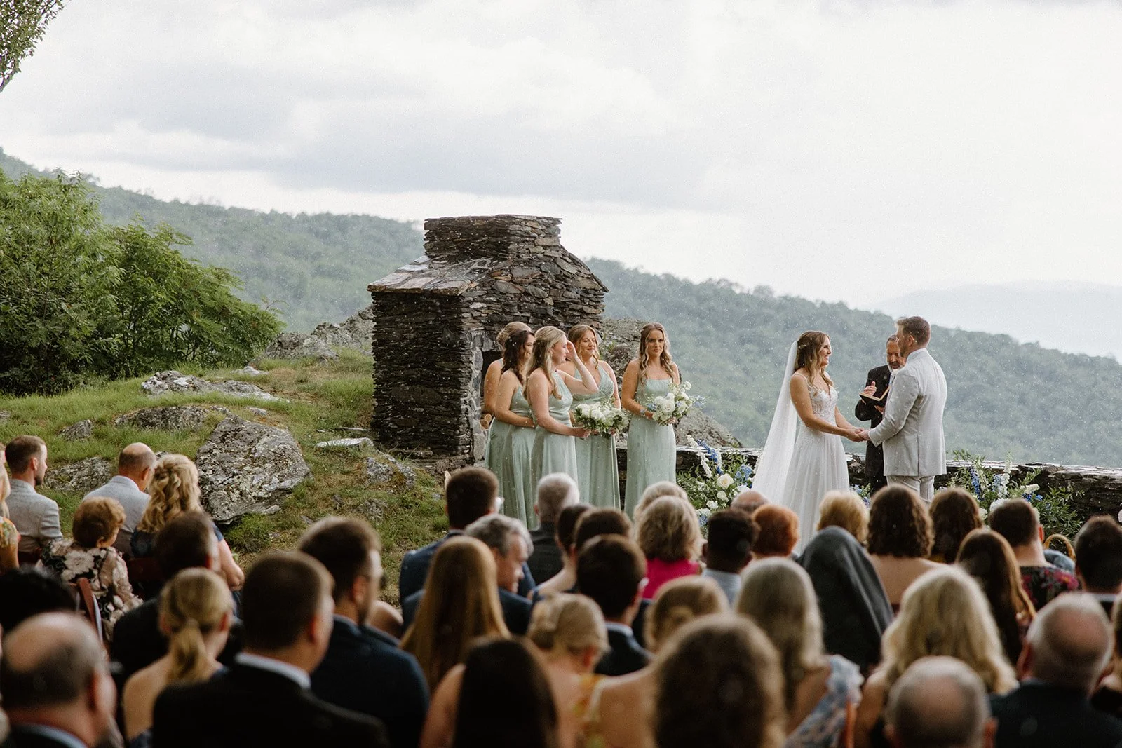 Outdoor mountain wedding ceremony on a stone terrace with guests seated and bridesmaids in blue dresses.