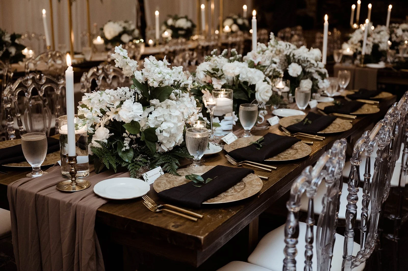 Elegant reception table with white floral centerpieces, taper candles, black napkins, and clear ghost chairs.