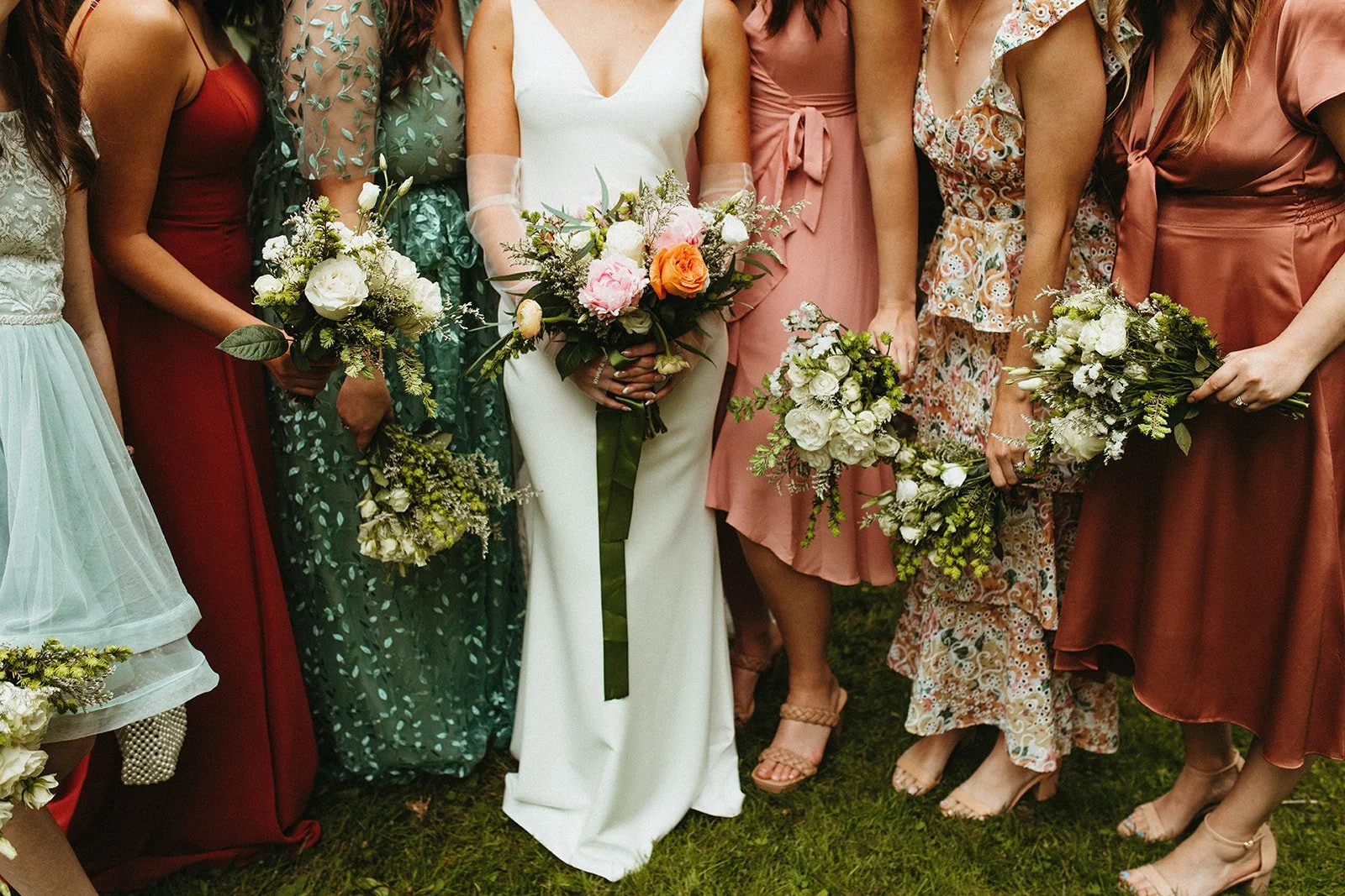 Cropped wedding party photo showing bridesmaids in mixed-color dresses holding bouquets, focused on flowers and lower half of outfits.