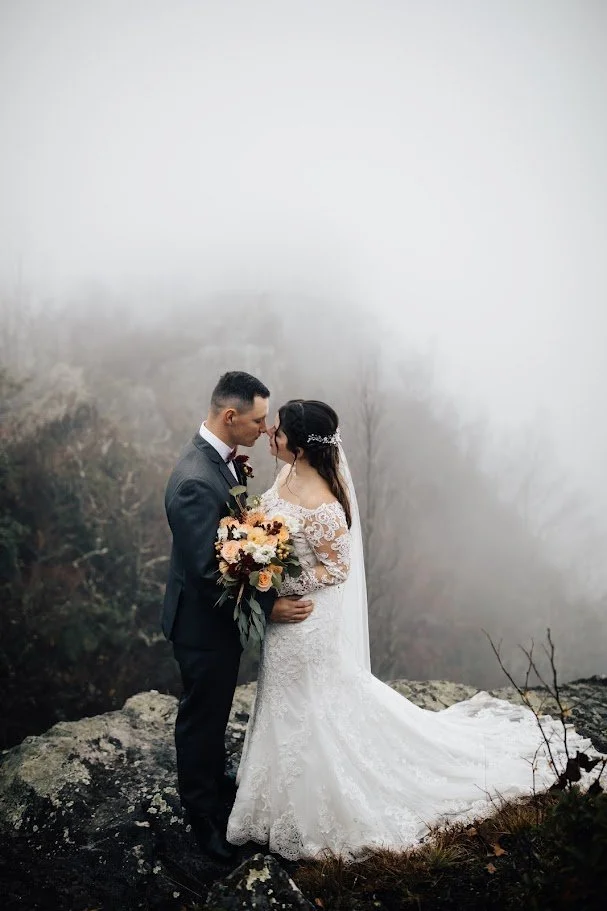Bride and groom embracing on a foggy cliffside with the bride’s long veil and train trailing behind.