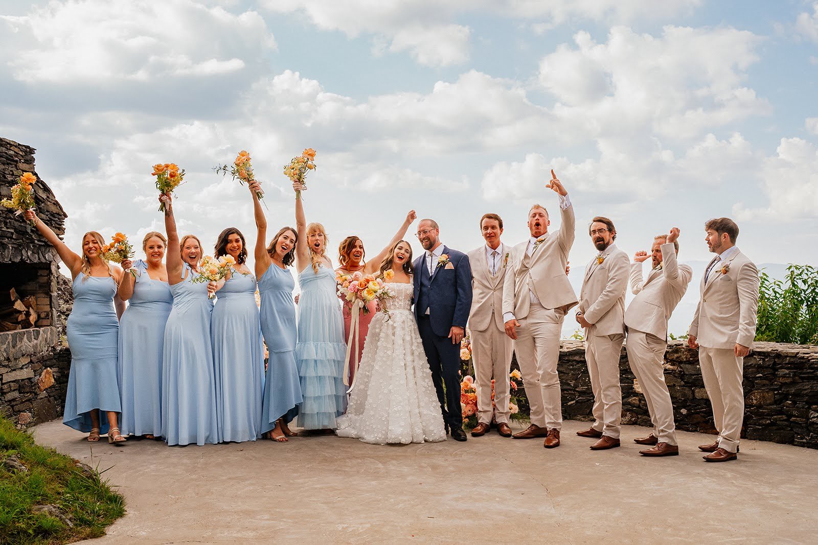 Wedding party celebrates outdoors with bridesmaids in light blue dresses and groomsmen in tan suits raising bouquets and hands.