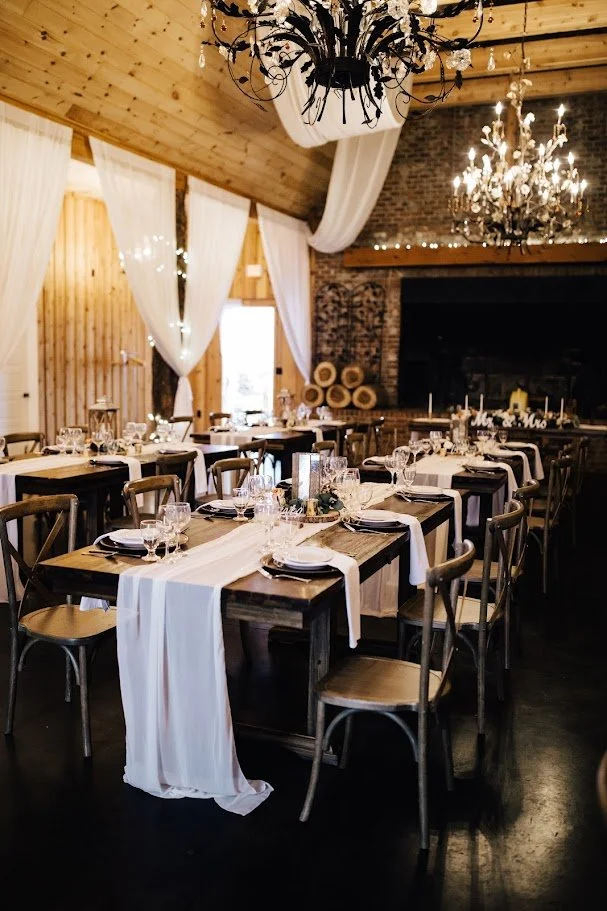 Rustic barn wedding reception with long tables, white draping, and chandeliers overhead.