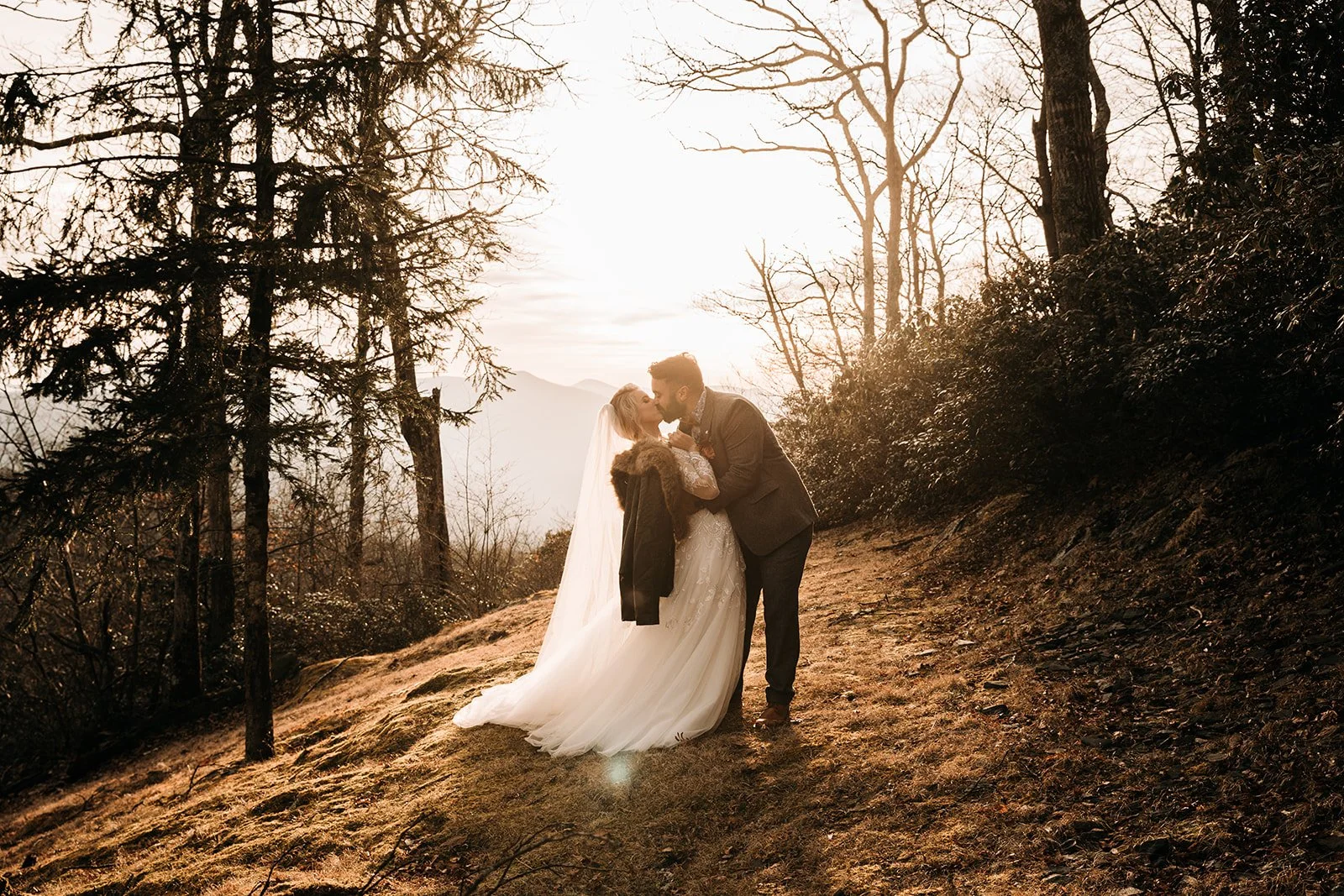 Bride and groom kissing at sunset on a wooded mountain slope with warm golden light behind them.