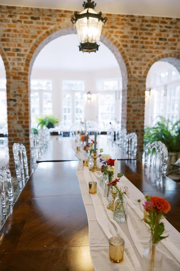 Long wooden reception table with floral bud vases and candles inside a brick-arched dining room.
