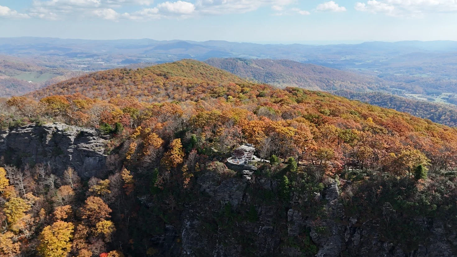 Aerial view of a rocky cliffside overlook surrounded by vibrant autumn trees and distant rolling mountains.