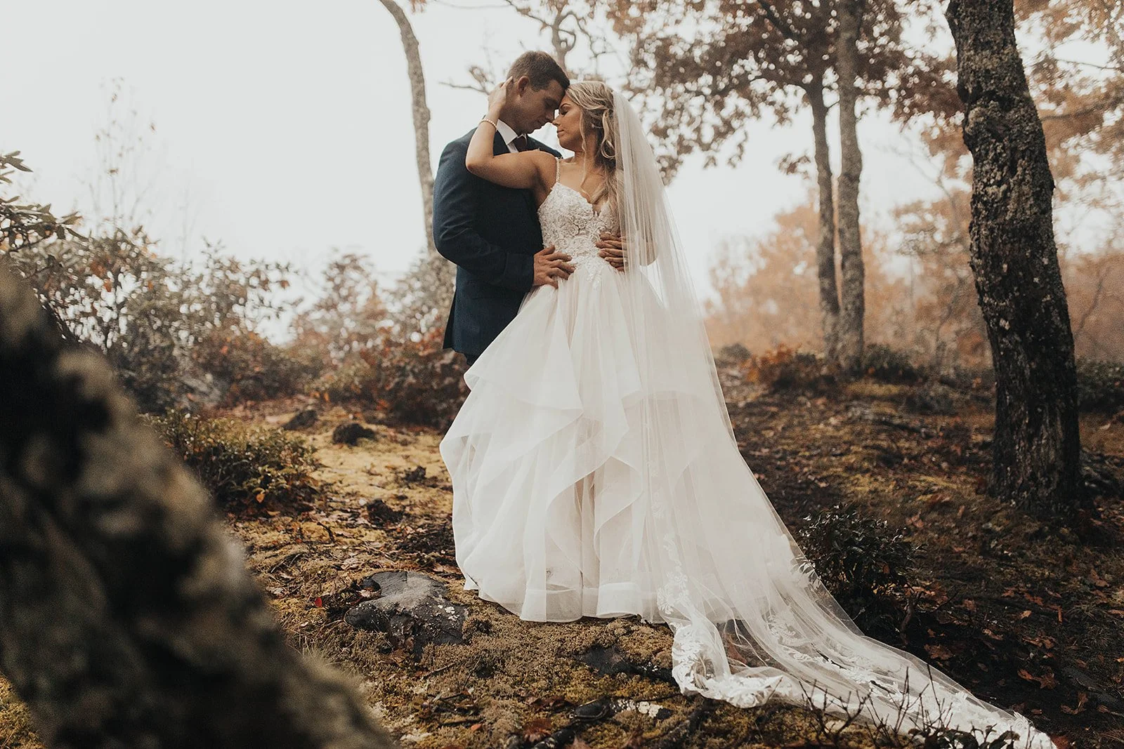 Bride and groom embracing in a wooded setting, with the bride’s long veil and flowing skirt draped over the ground in soft fall light