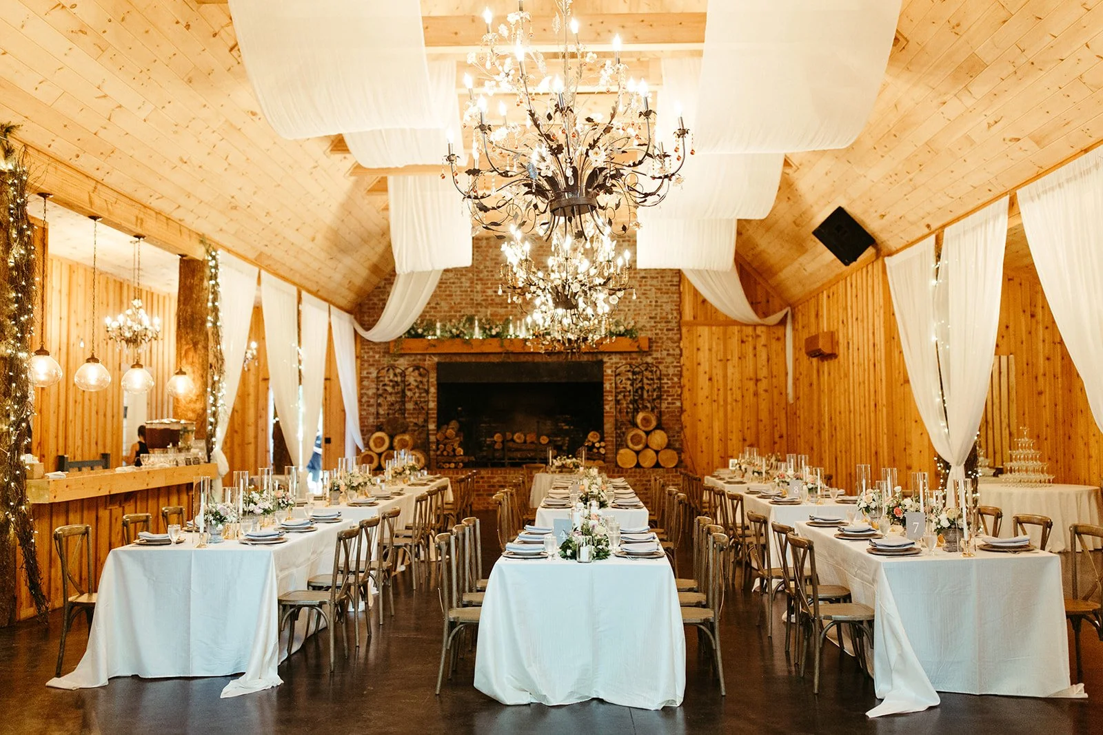 Rustic barn wedding reception with long tables, white ceiling draping, and chandeliers facing a fireplace.
