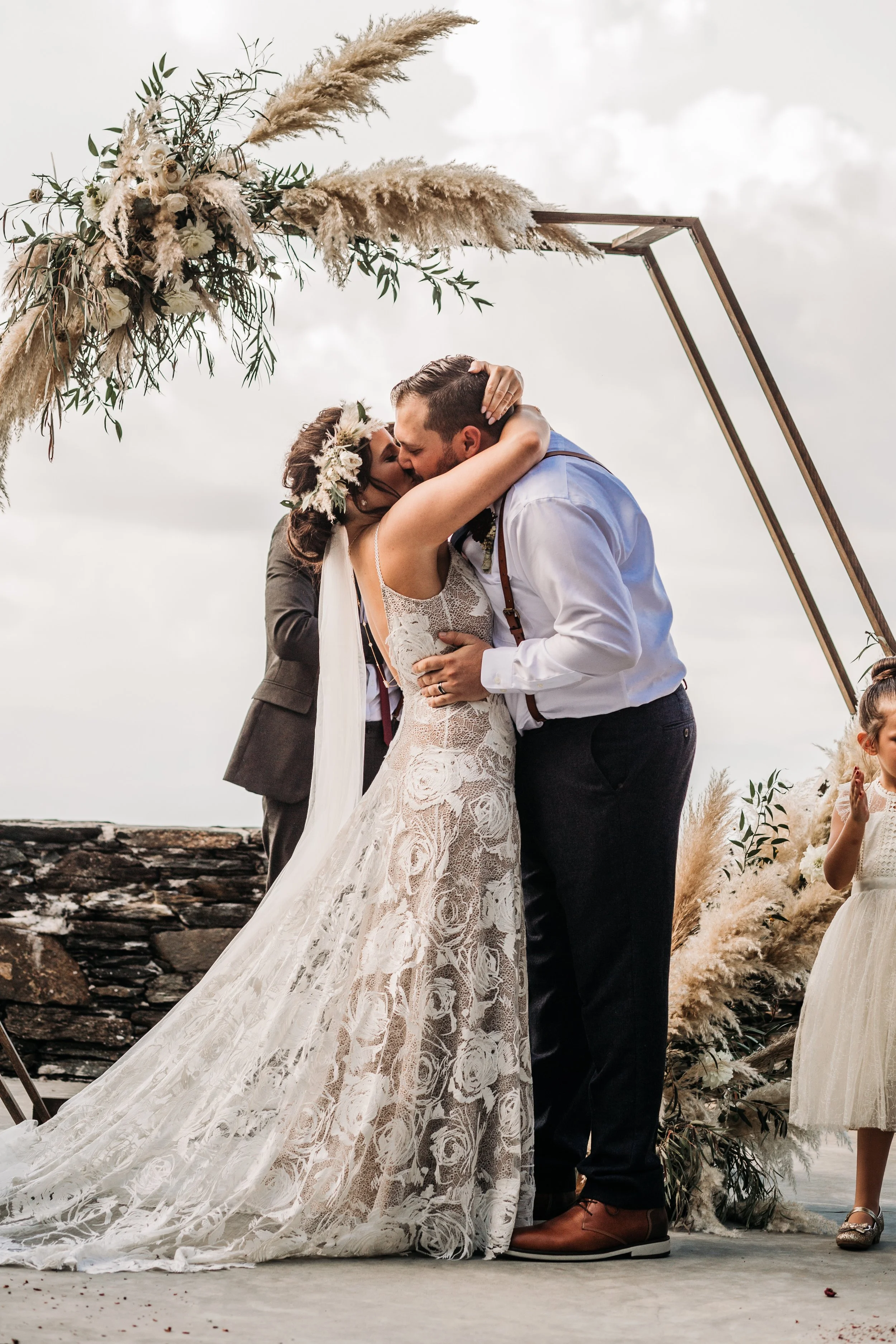 Ceremony first kiss under a geometric arch with pampas grass at a mountain overlook.