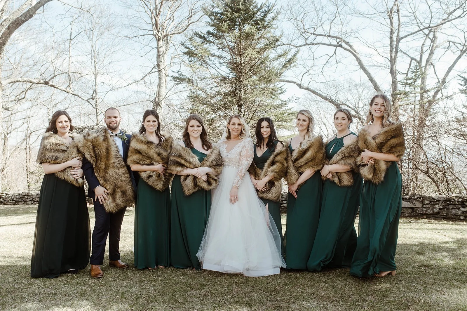 Bride standing with bridesmaids in emerald green dresses and fur wraps during outdoor wedding portraits.