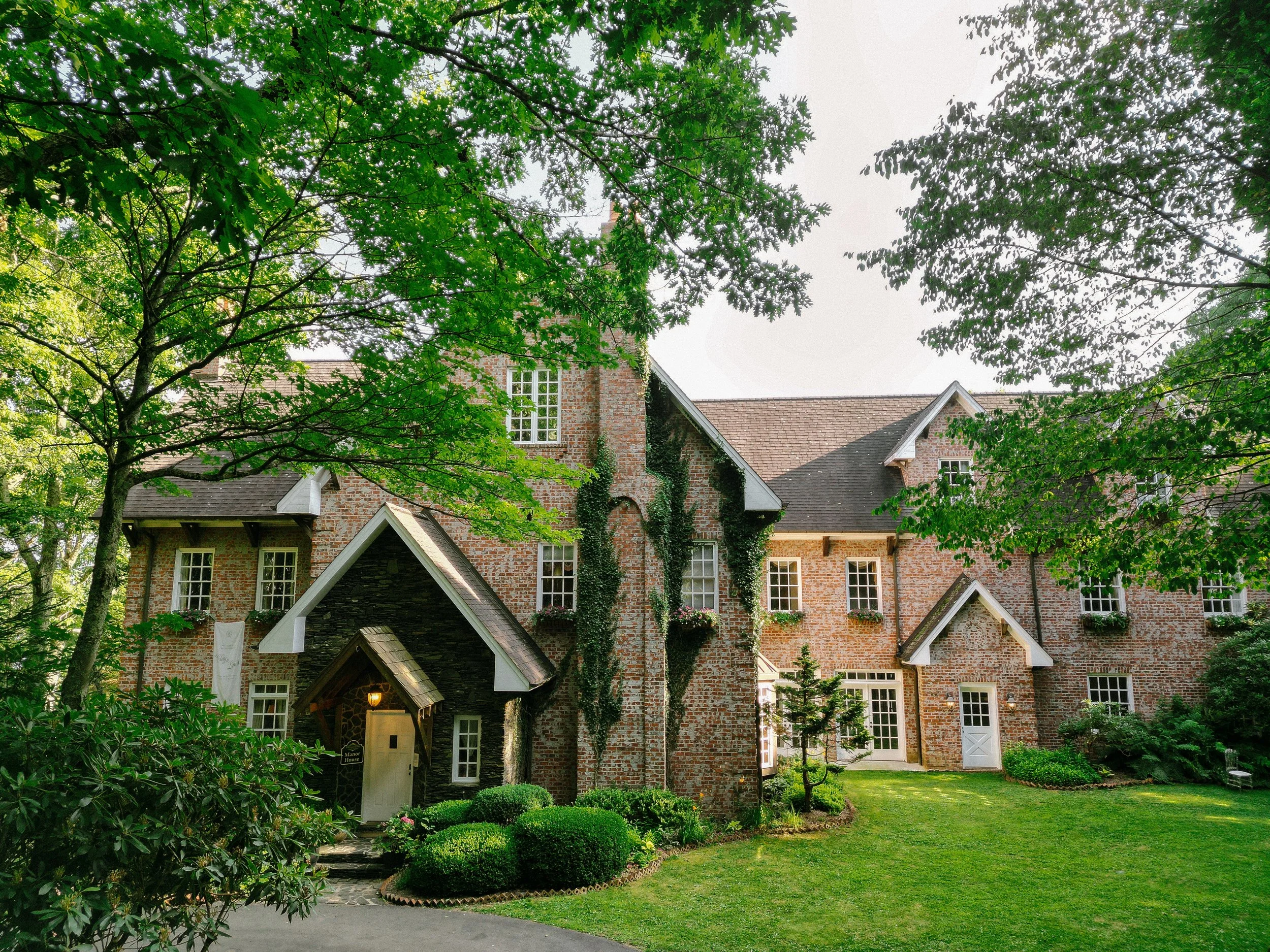 Historic brick estate wedding venue with ivy-covered walls, gabled rooflines, large windows, trees, and a green lawn.