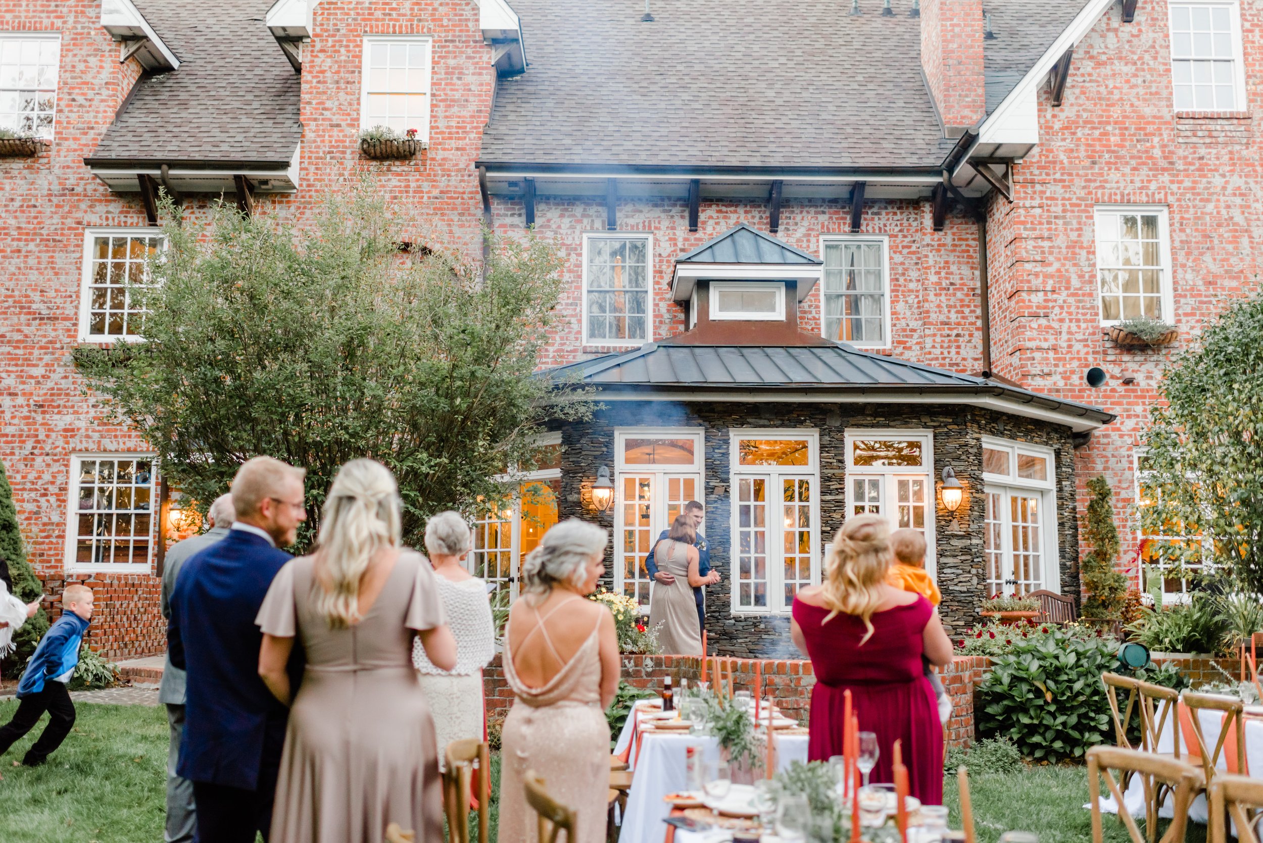 Outdoor wedding reception on the lawn outside a historic brick estate with guests, dinner tables, and warm window lighting.