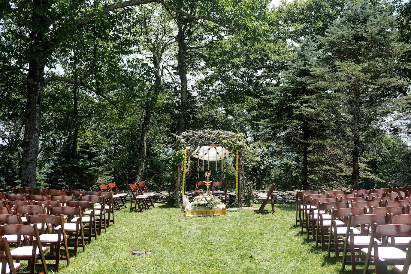 Rows of chairs face a decorated ceremony canopy on a grassy lawn surrounded by trees in a quiet outdoor setting.