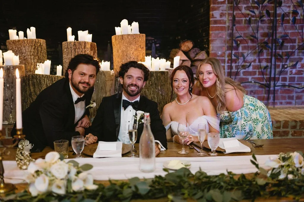 Bride and groom smiling with guests at a candlelit sweetheart table decorated with greenery, white flowers, and champagne glasses.
