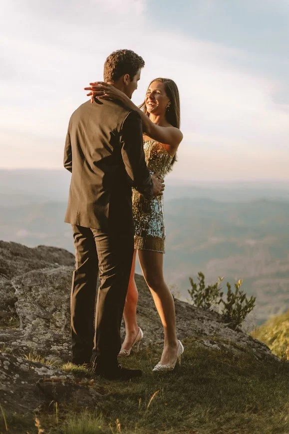 Couple embraces on a rocky overlook at sunset, with sweeping mountain views behind them.