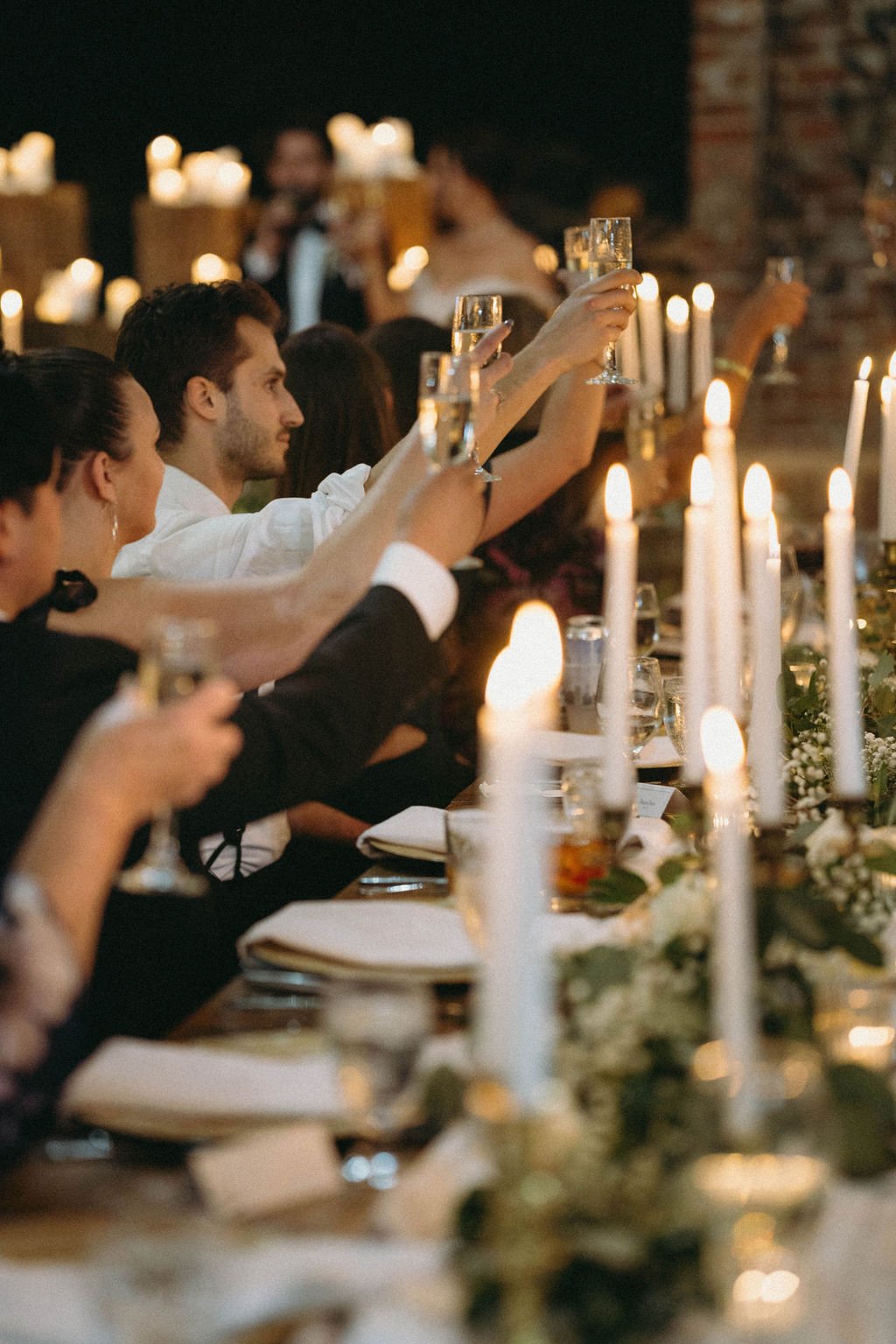 Wedding guests raising glasses for a toast at a candlelit reception dinner with greenery and white taper candles.
