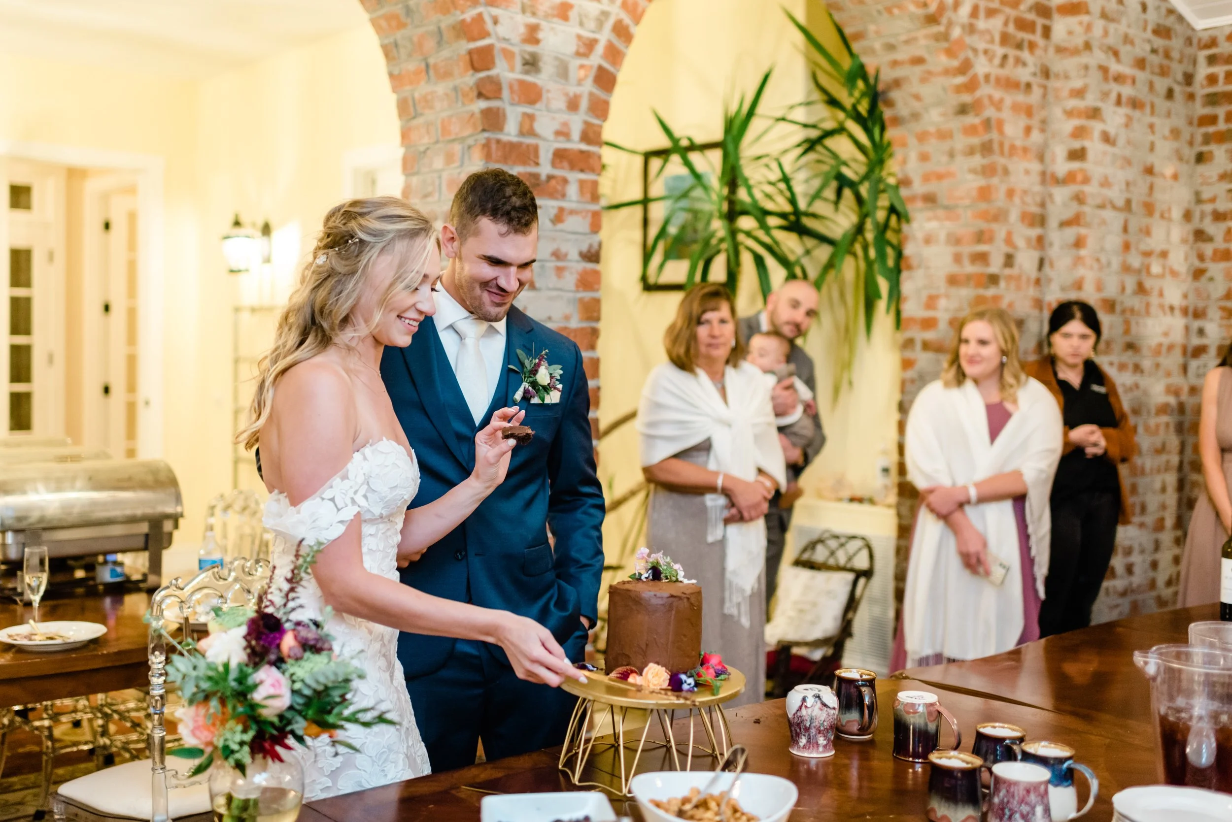 Bride and groom cutting a small chocolate wedding cake indoors near exposed brick walls and family members.