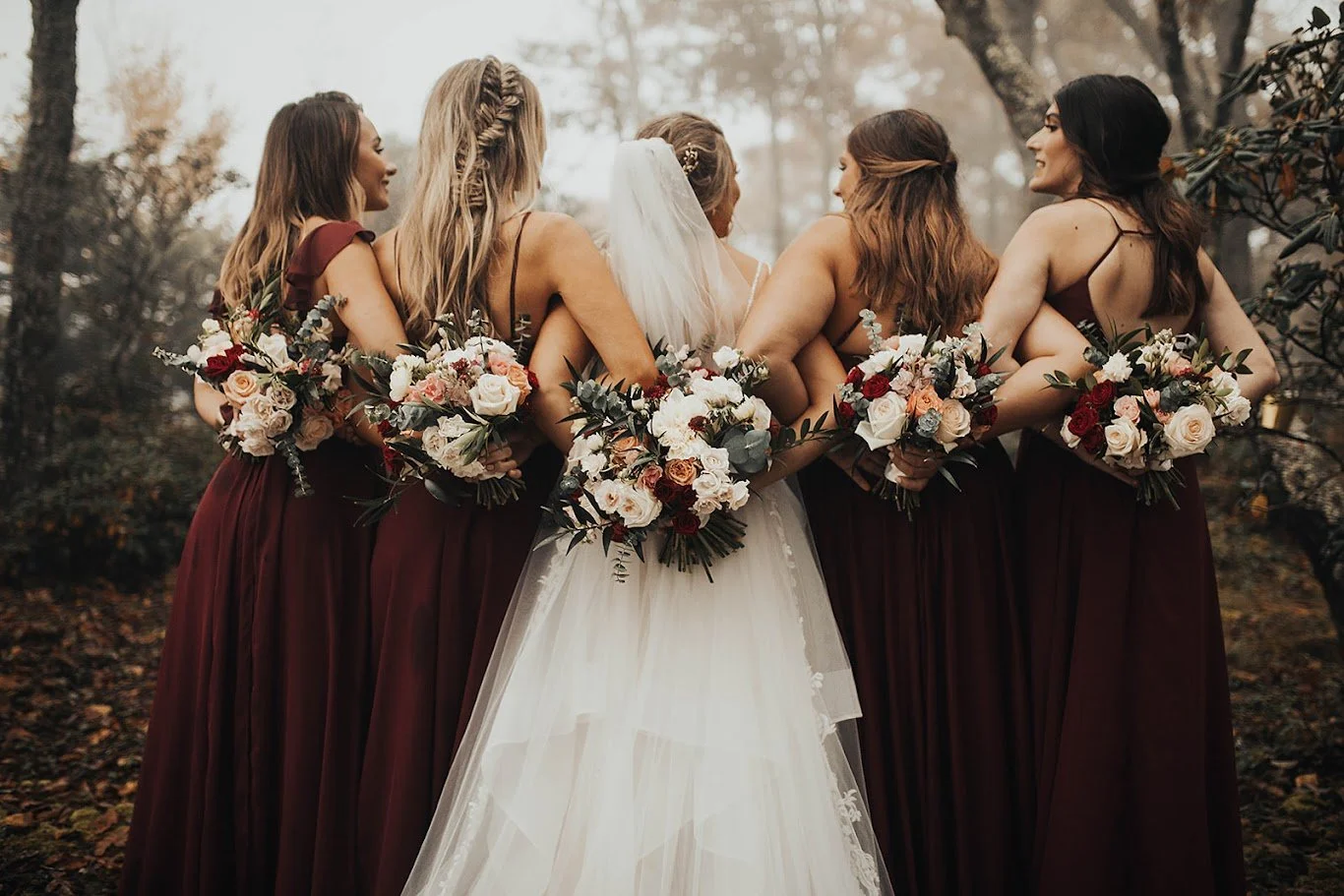 Bride with bridesmaids in burgundy dresses holding bouquets behind their backs in a foggy forest.