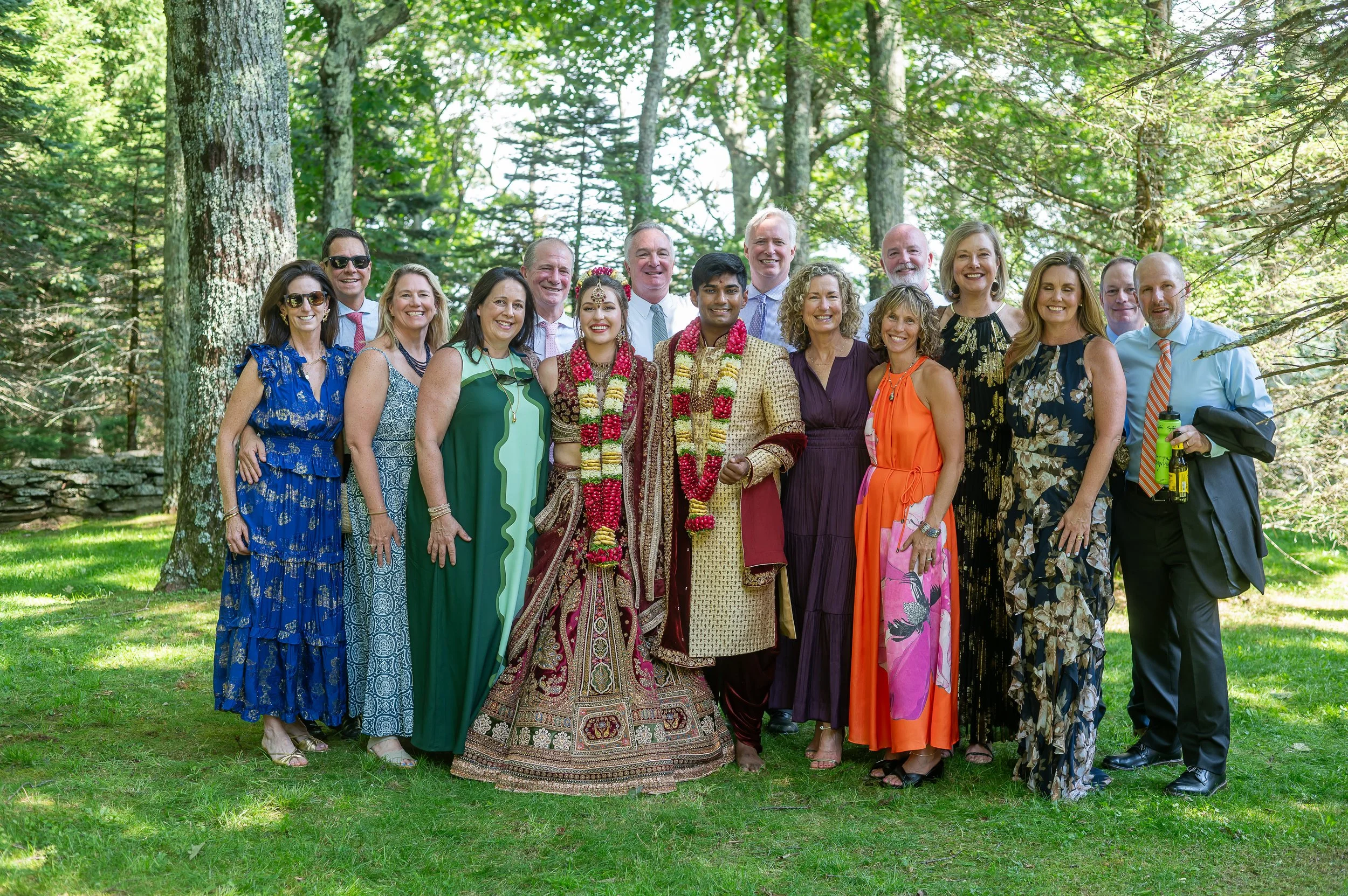 Outdoor group wedding portrait with the couple in traditional Indian attire standing with family and friends on a wooded lawn.