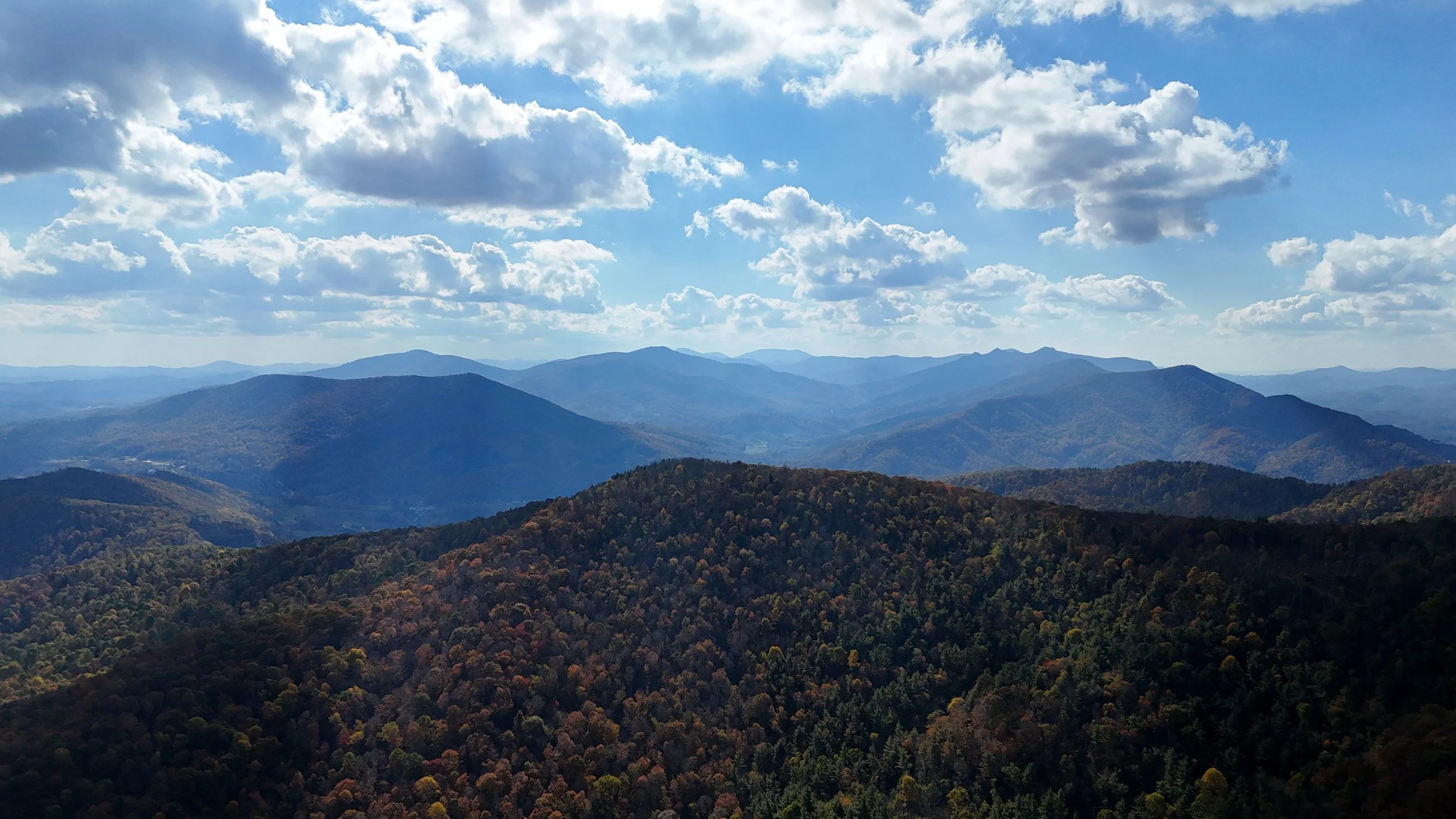 Panoramic view of layered blue mountains and a forested ridge in autumn colors beneath a sky filled with bright clouds.