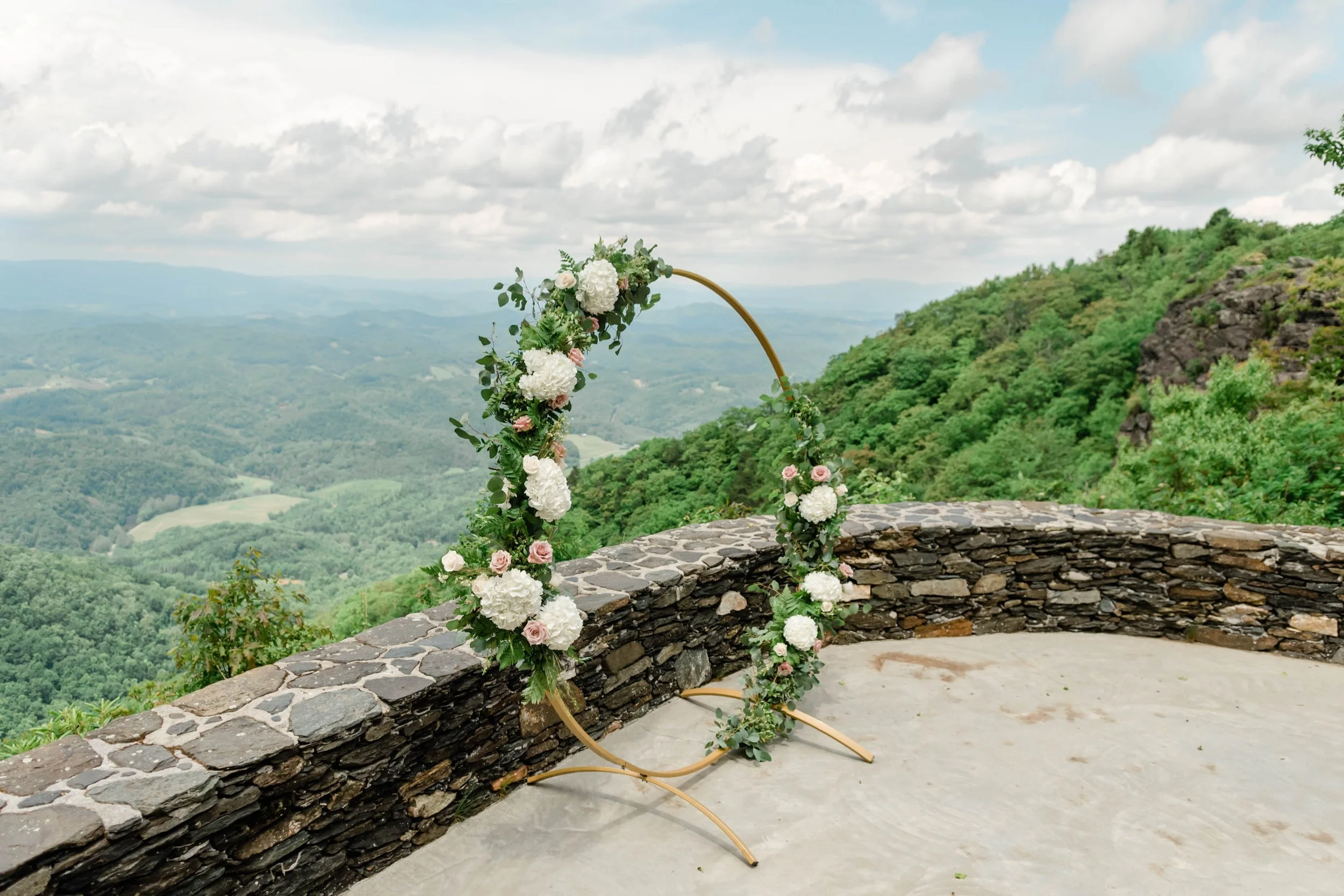 Circular wedding arch decorated with white flowers and greenery on a stone terrace overlooking the mountains