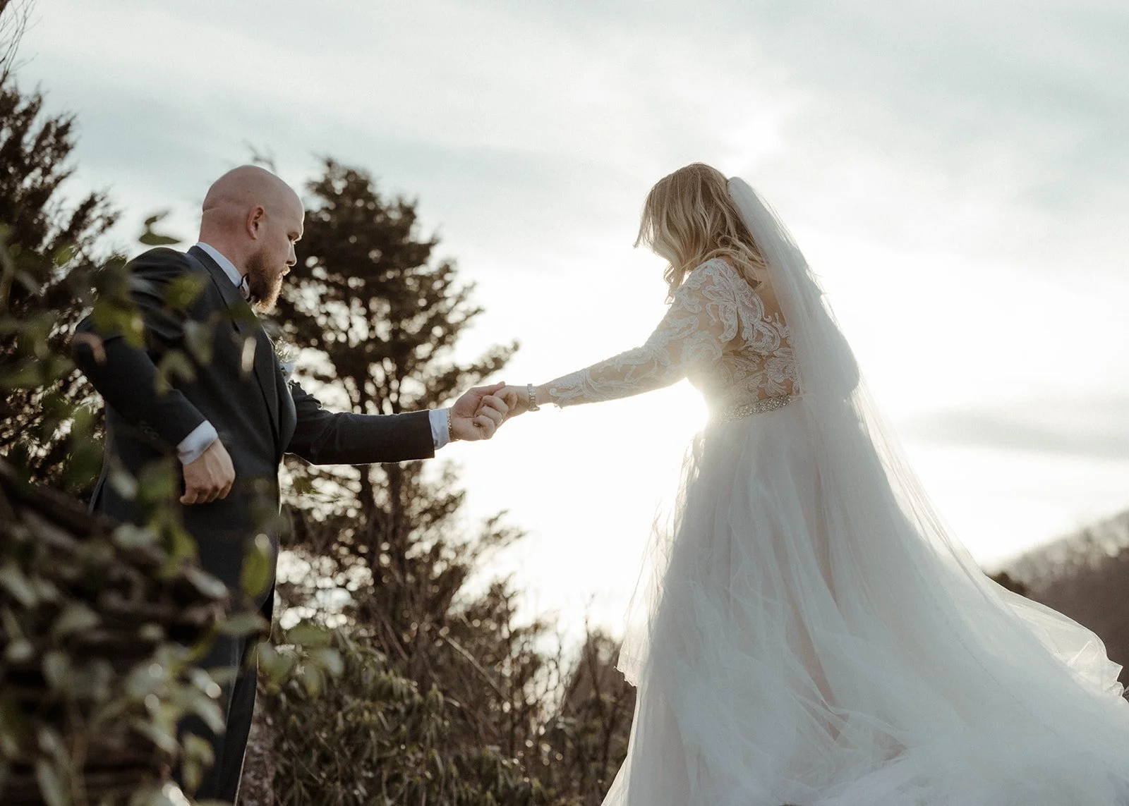 Bride reaching for the groom’s hand during a romantic backlit mountain wedding portrait.