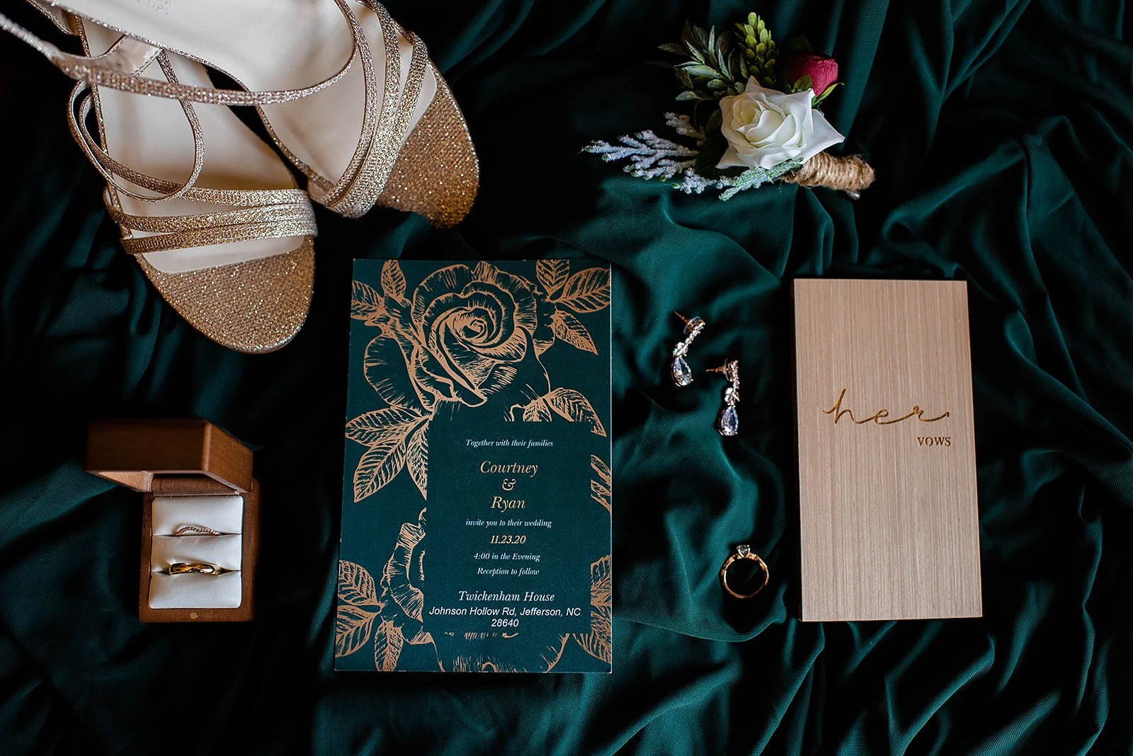 Wedding invitation flat lay on emerald fabric with gold shoes, rings, earrings, boutonniere, and vow books.