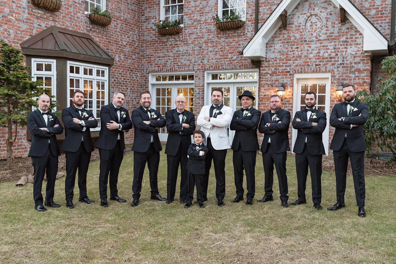 Groomsmen group portrait outside a brick venue, one wearing a white dinner jacket.