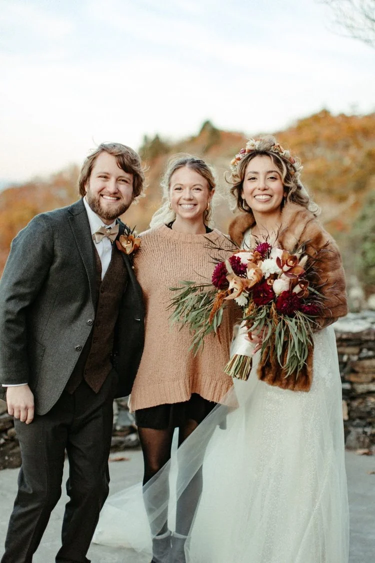 Smiling bride in a fur stole holding a bouquet, groom in a suit, and a friend in a sweater pose together at a North Carolina mountain wedding.