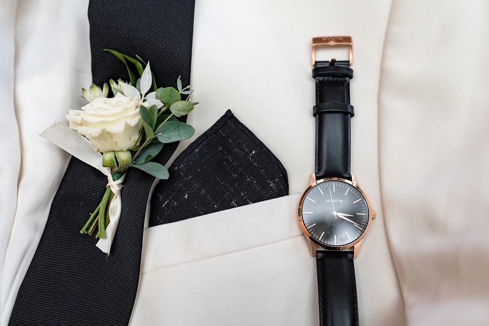 Groom accessories flat lay with watch, boutonniere, and pocket square on light fabric.