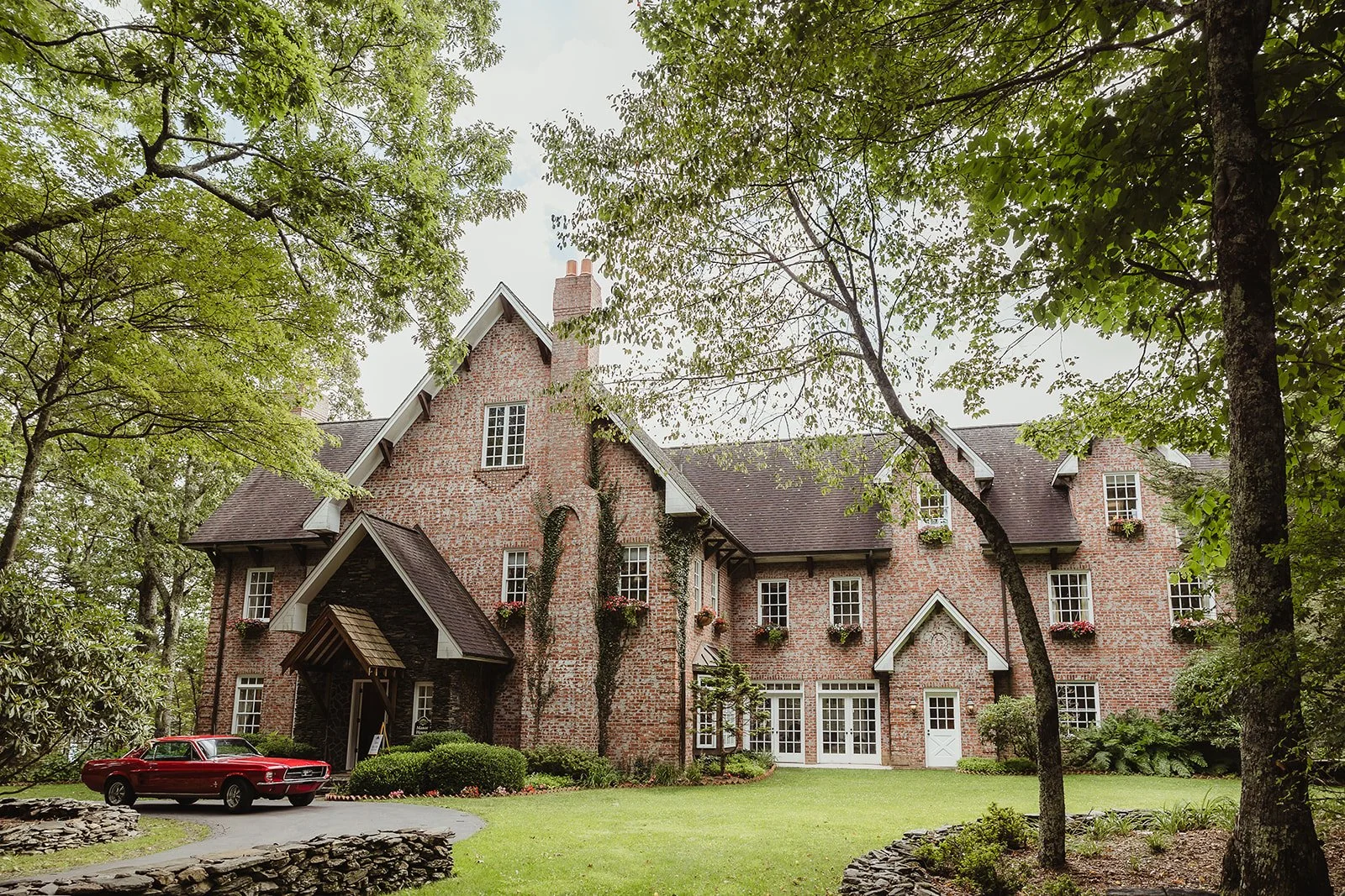 Large brick estate wedding venue surrounded by trees and a green lawn, viewed from the driveway.