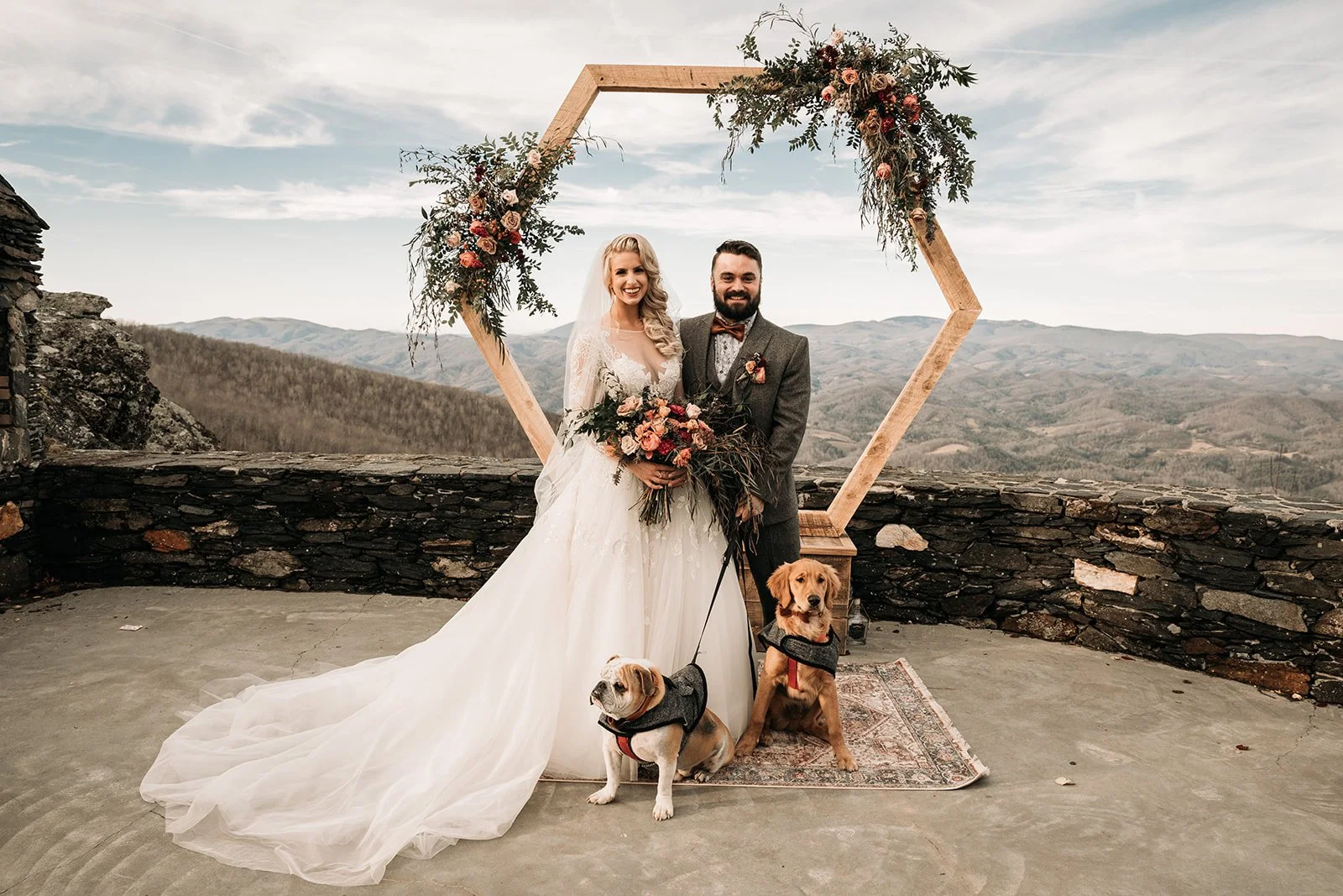 Bride and groom posing with their dogs beneath a hexagon floral wedding arch overlooking the mountains.