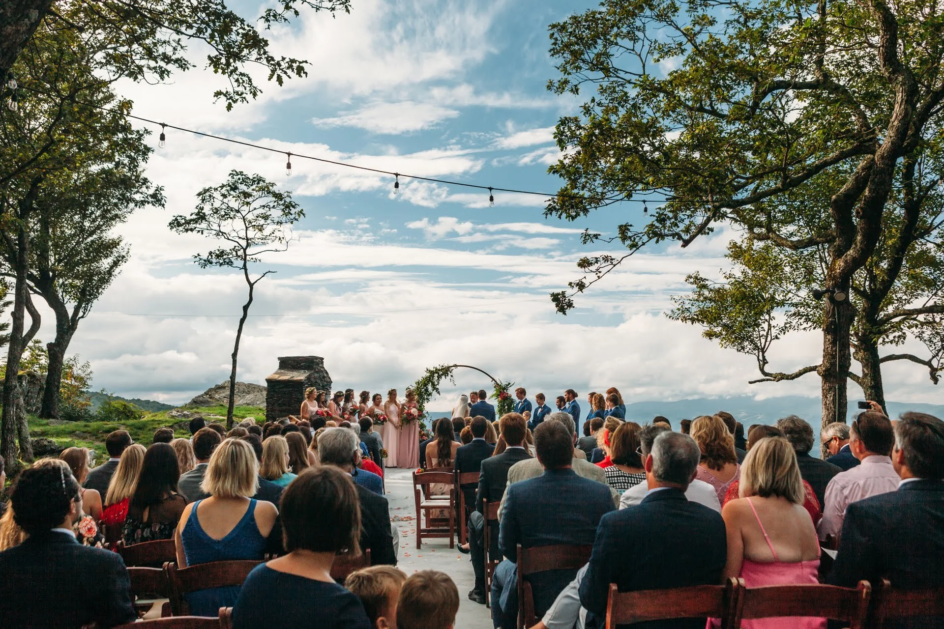Outdoor wedding ceremony at a mountain overlook with string lights and guests seated.