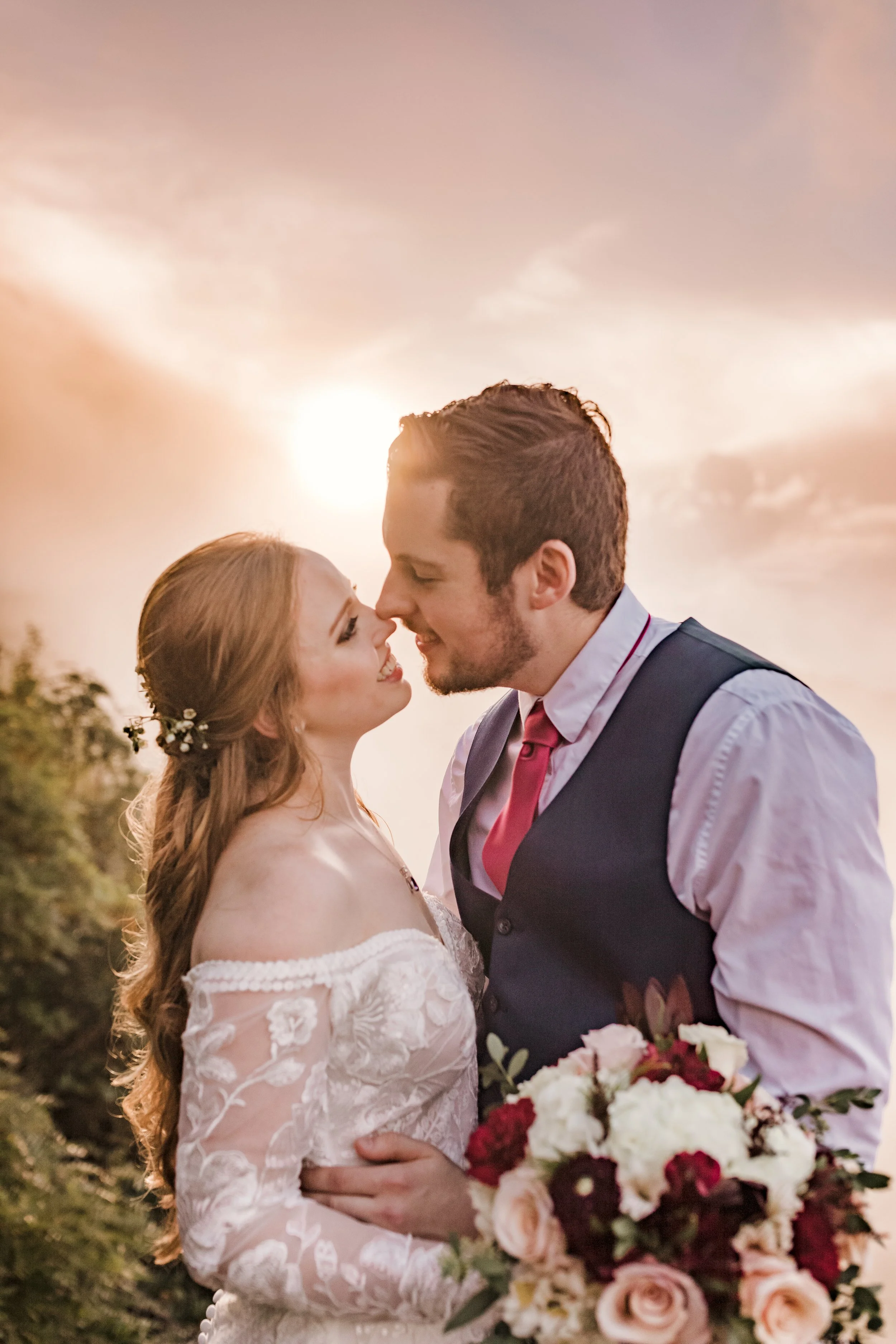 A bride in white and groom in vest kiss on a rocky mountain, with forested peaks and cloudy sky.