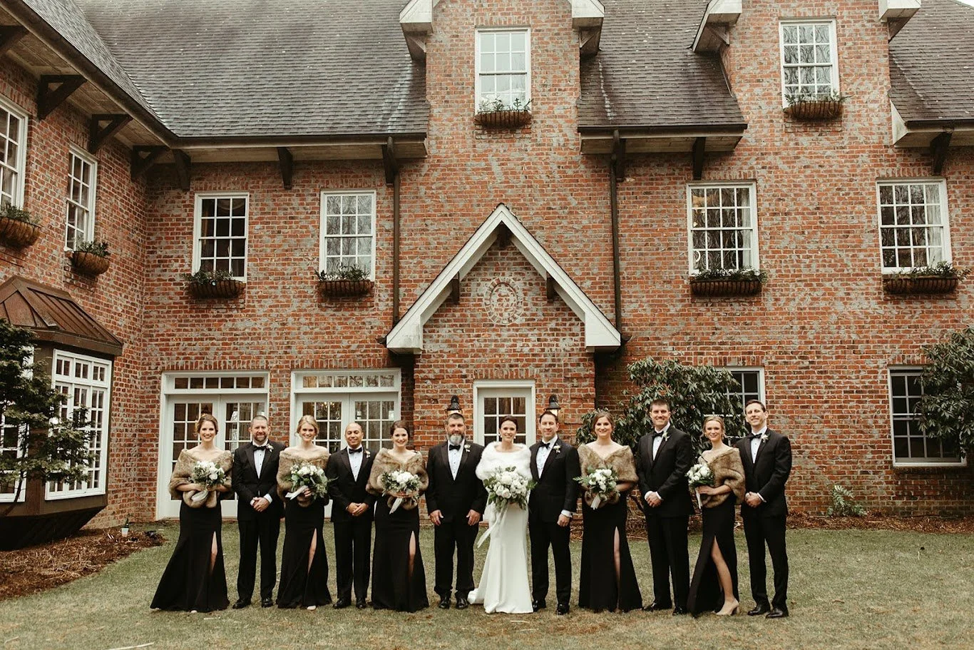 Full wedding party group portrait in front of a brick venue, dressed in black tie attire.