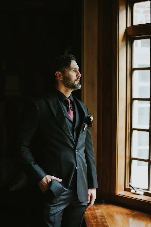 Groom in a dark suit with a red tie standing by a window in natural light.