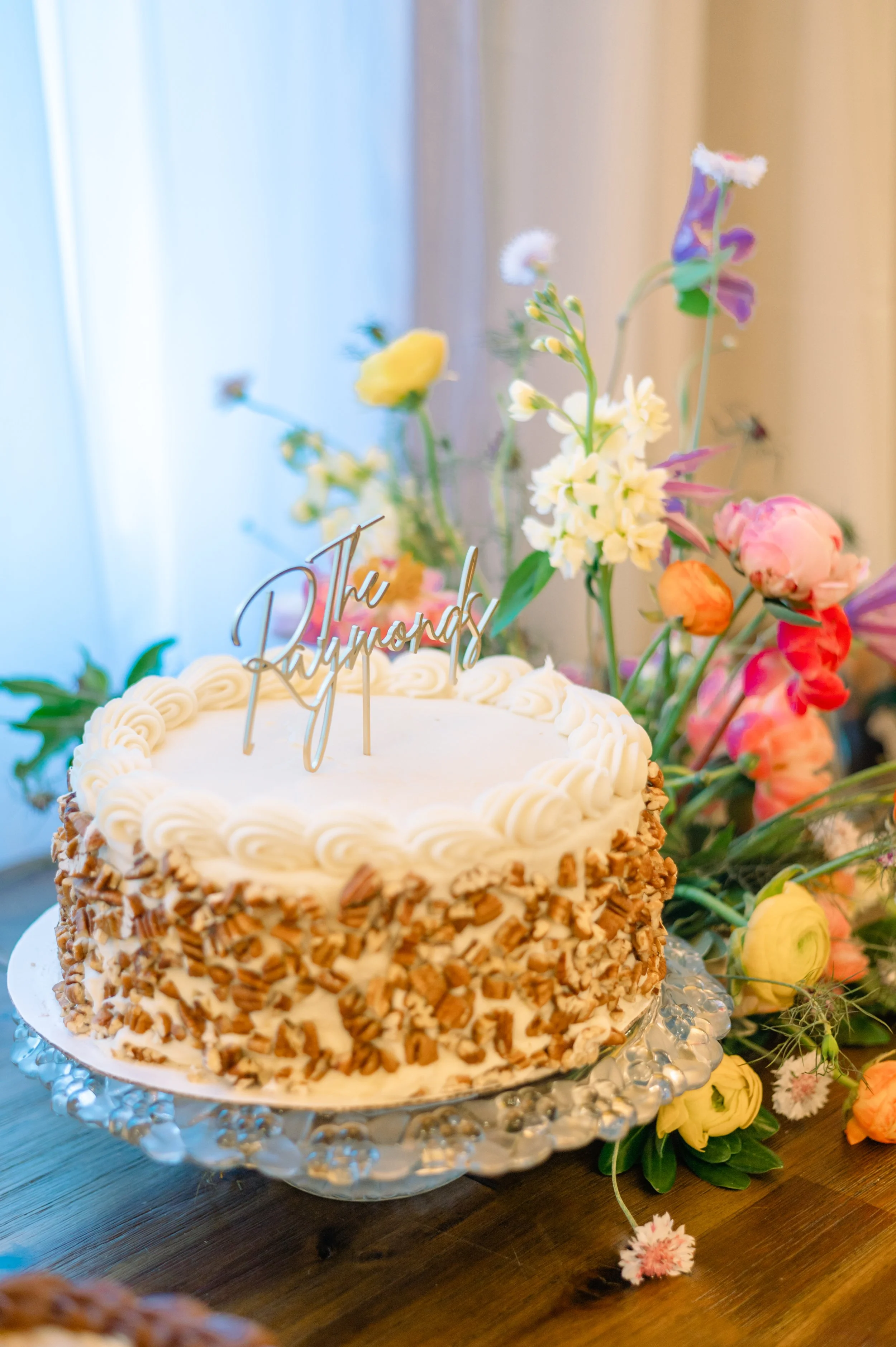 Single-tier wedding cake with piped frosting on a glass stand beside colorful flowers