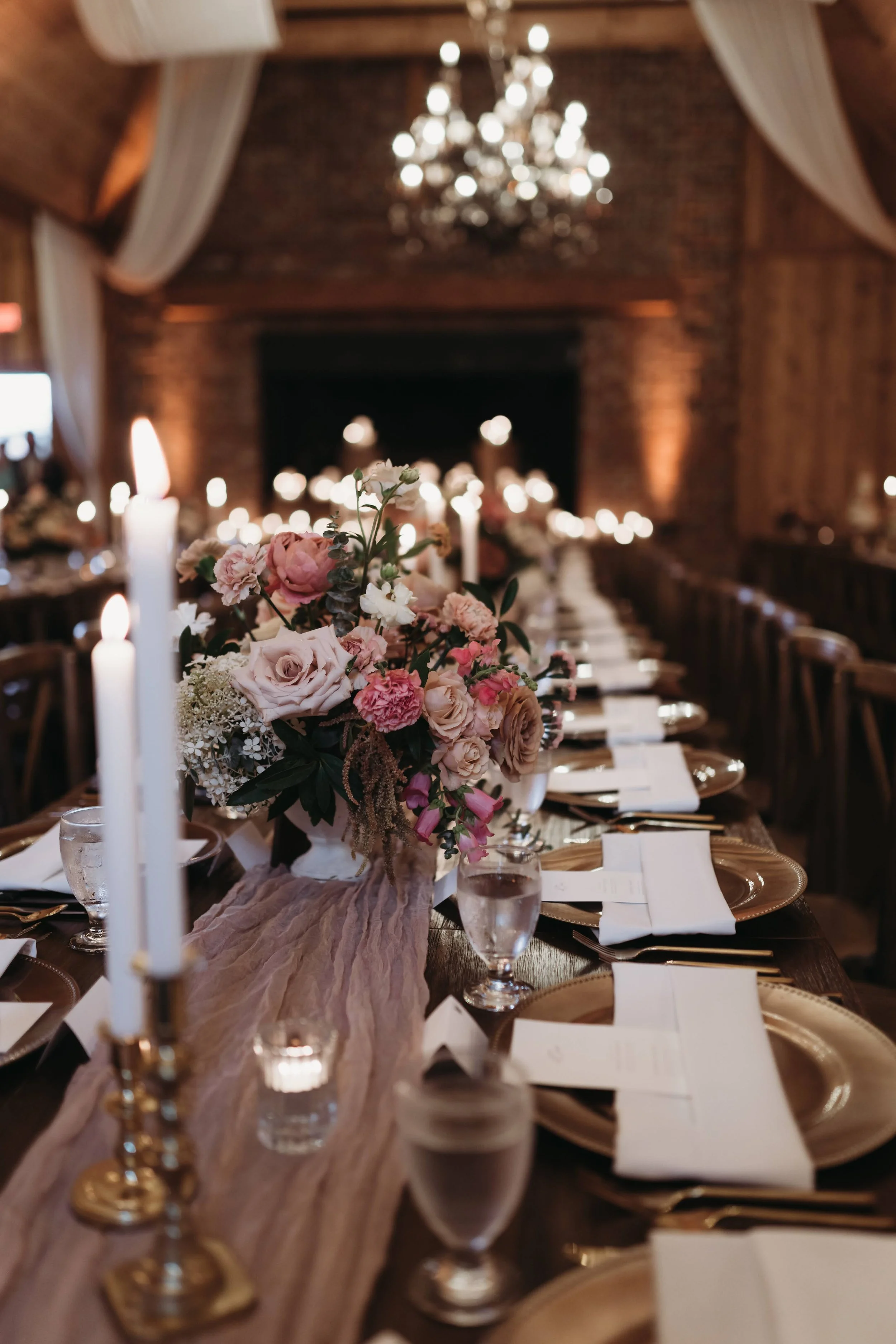 Candlelit wedding reception table with blush floral centerpieces, gold place settings, white draping, and chandelier lighting.