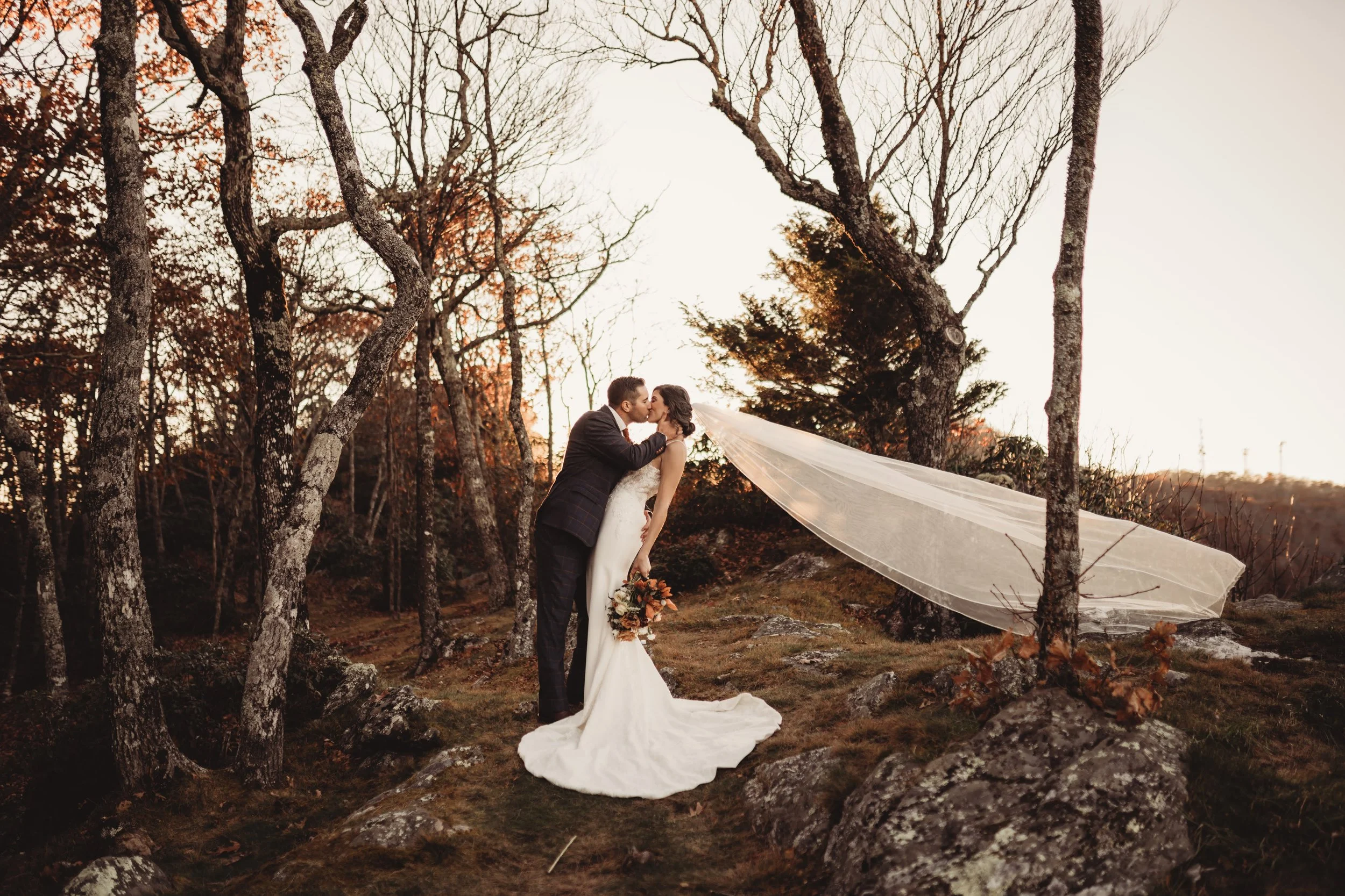 Bride and groom kissing in a forest as the bride’s veil blows dramatically to the side