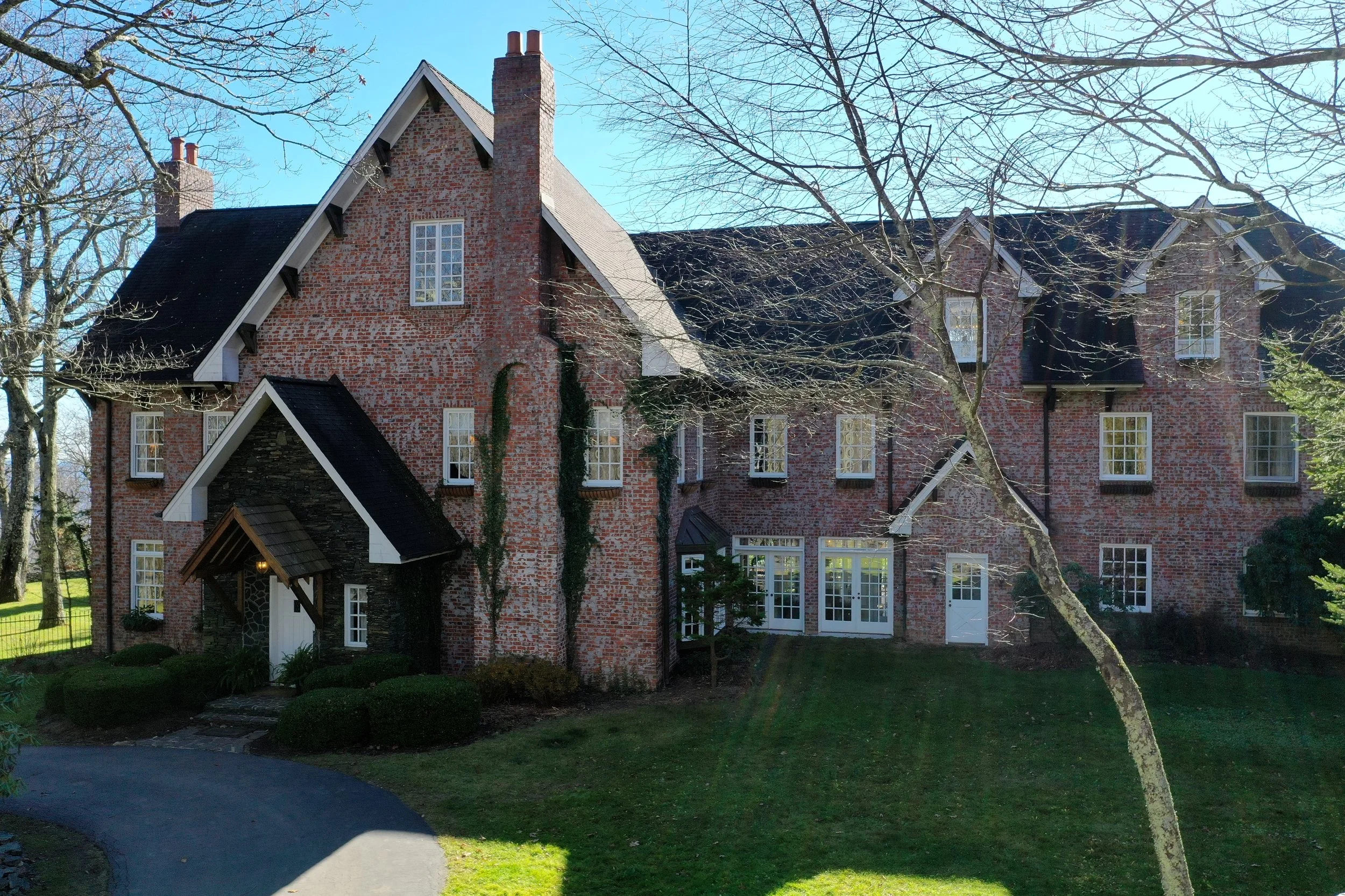 Front exterior of a brick estate home with gables, chimneys, and a green lawn on a sunny day