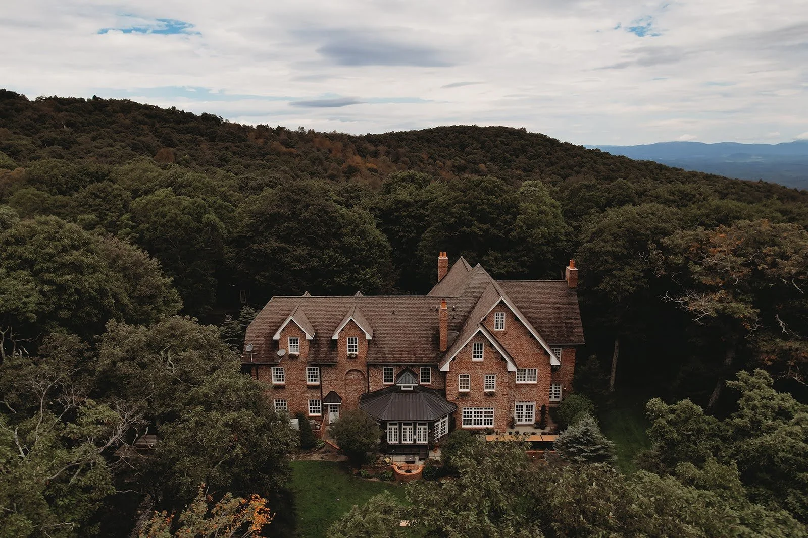 Aerial view of a large brick manor house surrounded by dense forest with rolling mountains under a cloudy sky.