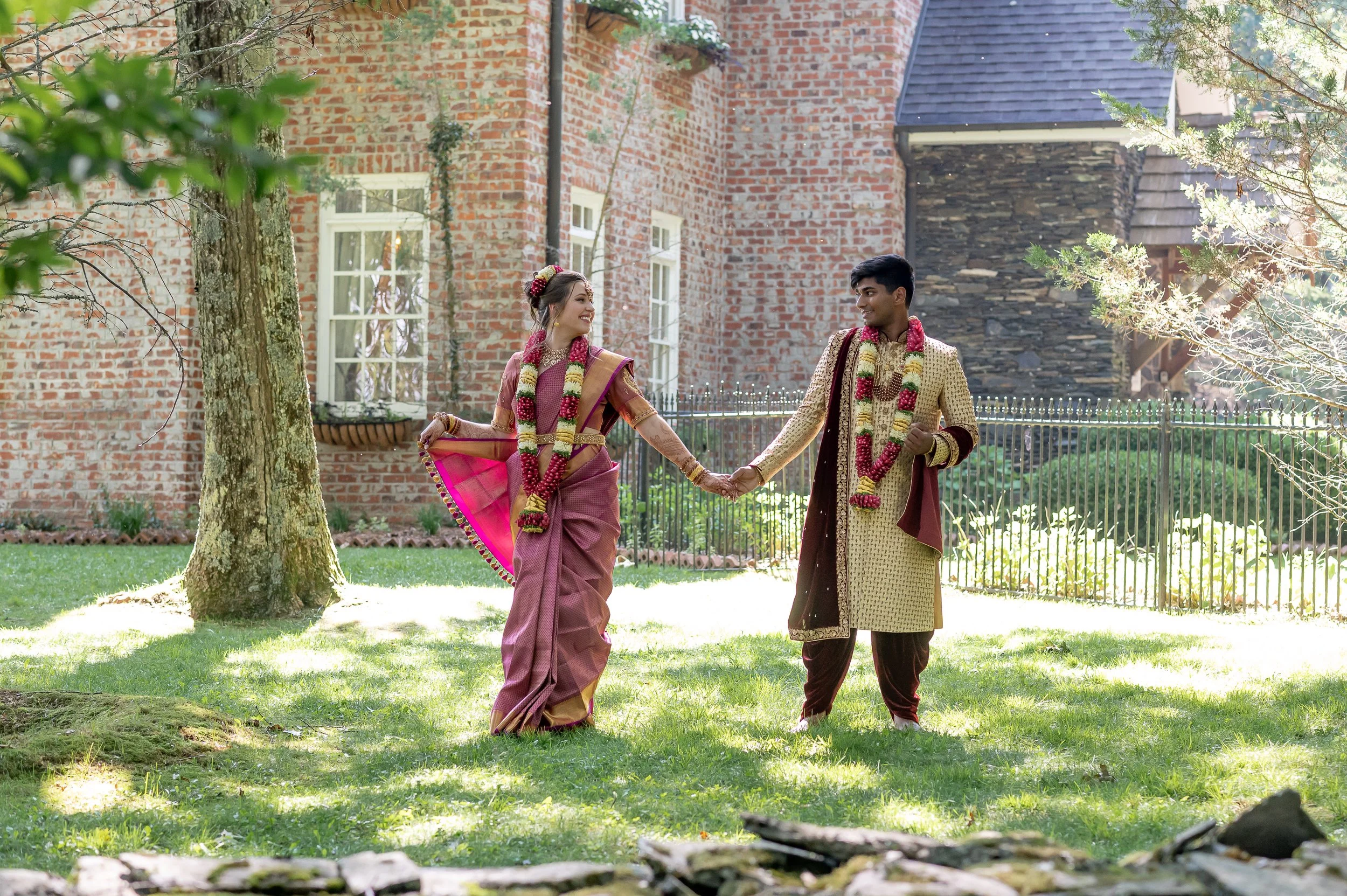 Couple in traditional South Asian clothing and flower garlands hold hands on a sunny lawn beside a brick building.