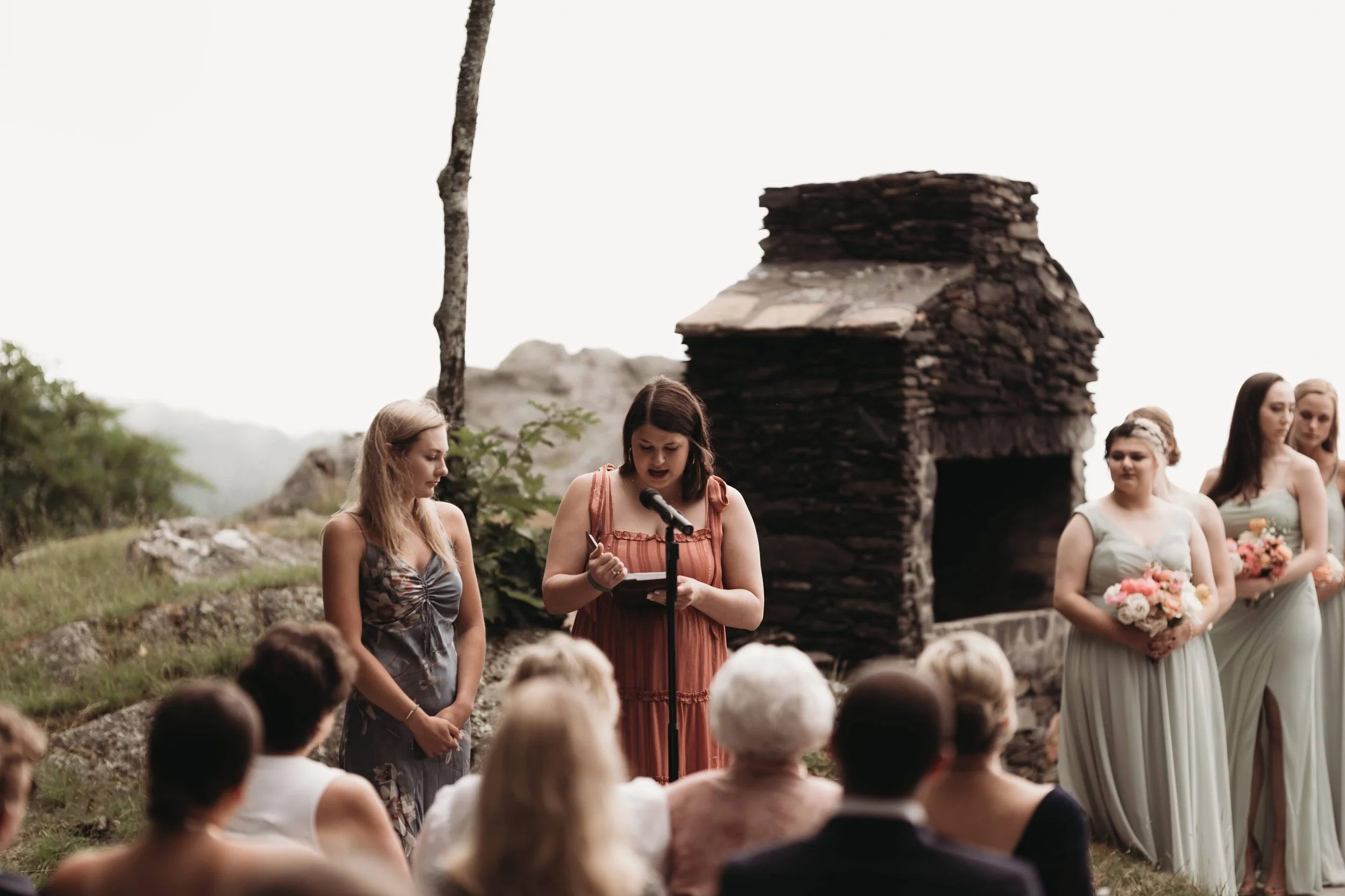 Outdoor wedding ceremony where a woman reads at a microphone while bridesmaids stand nearby near a small stone structure.