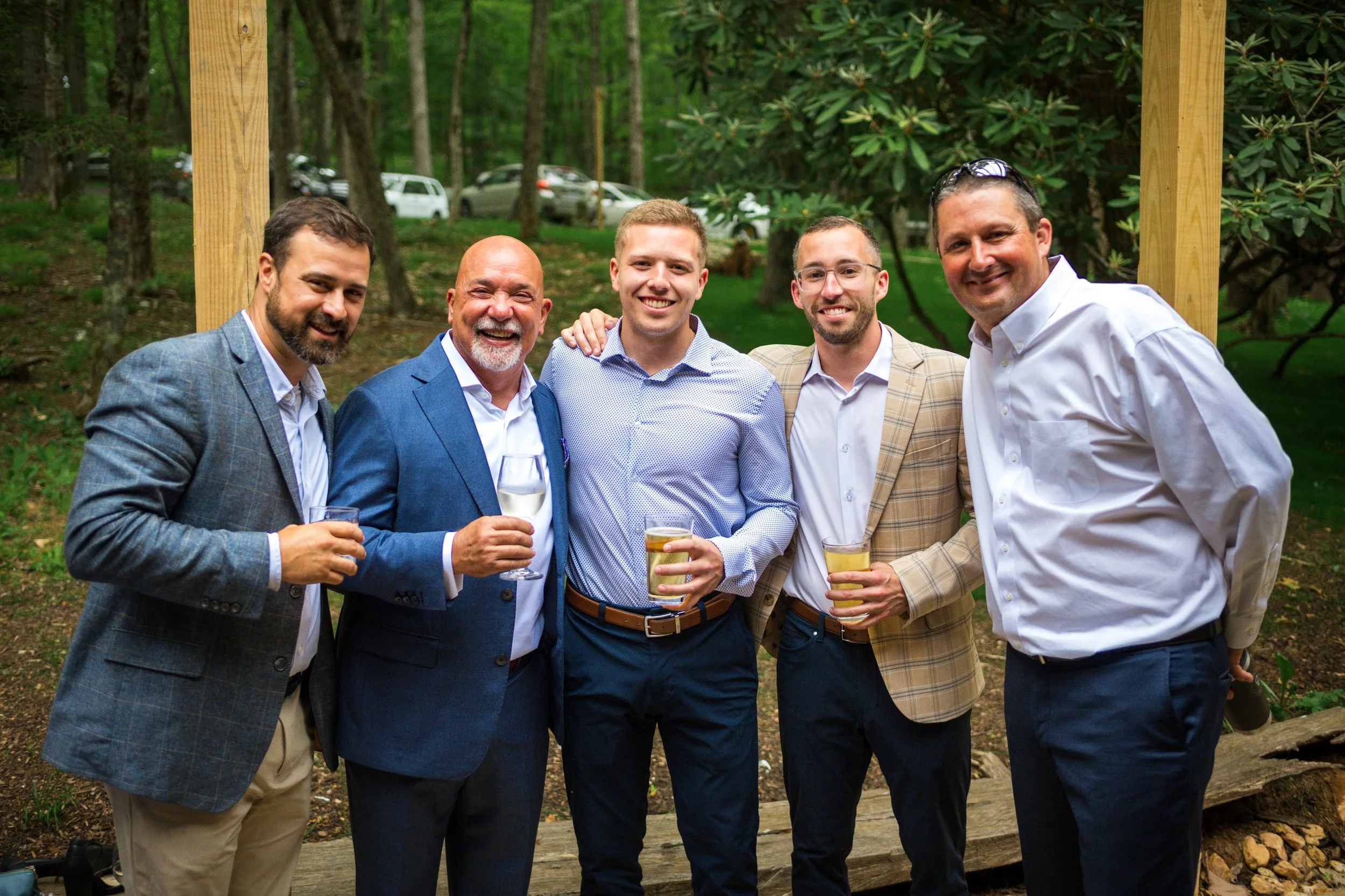 Business retreat attendees smiling and networking with drinks in a wooded outdoor venue setting.