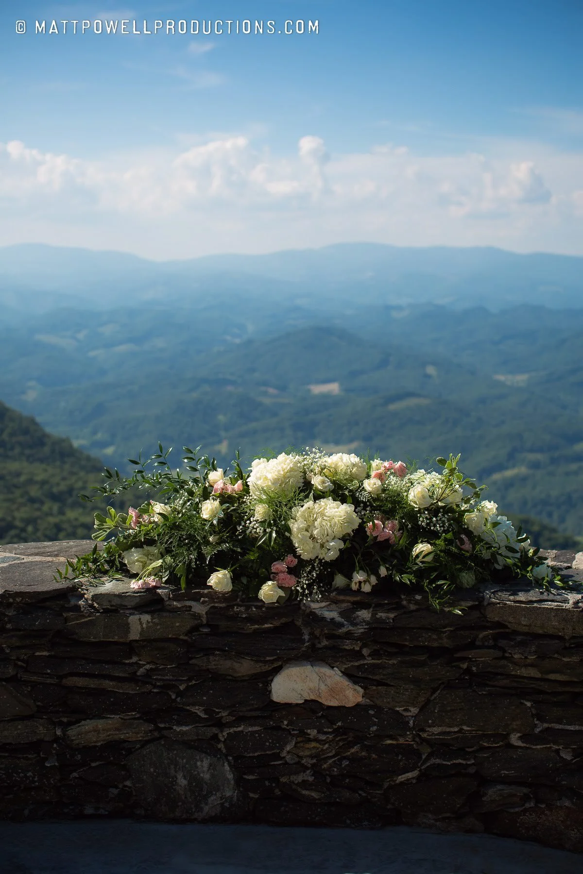Wedding floral arrangement on a stone wall with blue mountain views beyond.