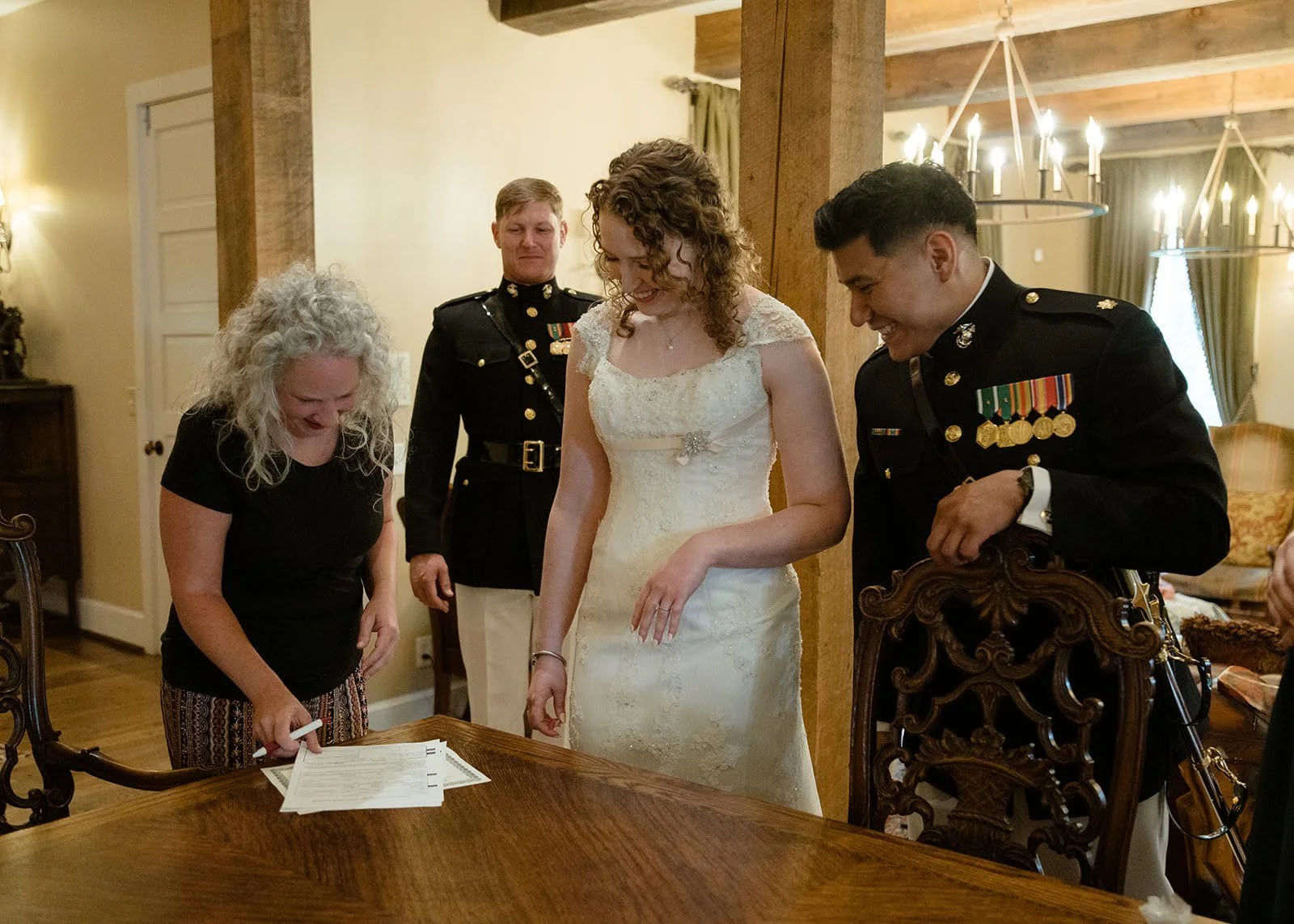 Bride and military grooms signing wedding documents at a wooden table inside a rustic venue.