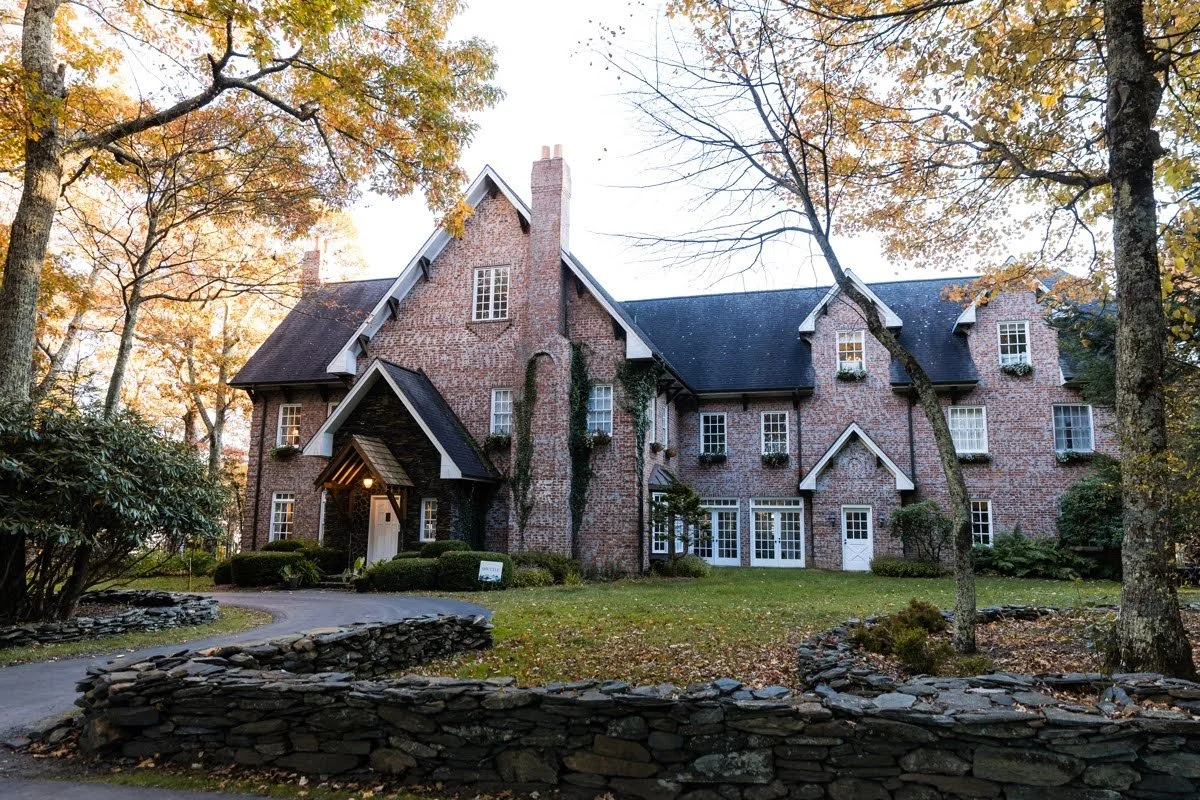 Brick inn-style building in autumn with trees overhead, a curved driveway, and a low stone wall in front.